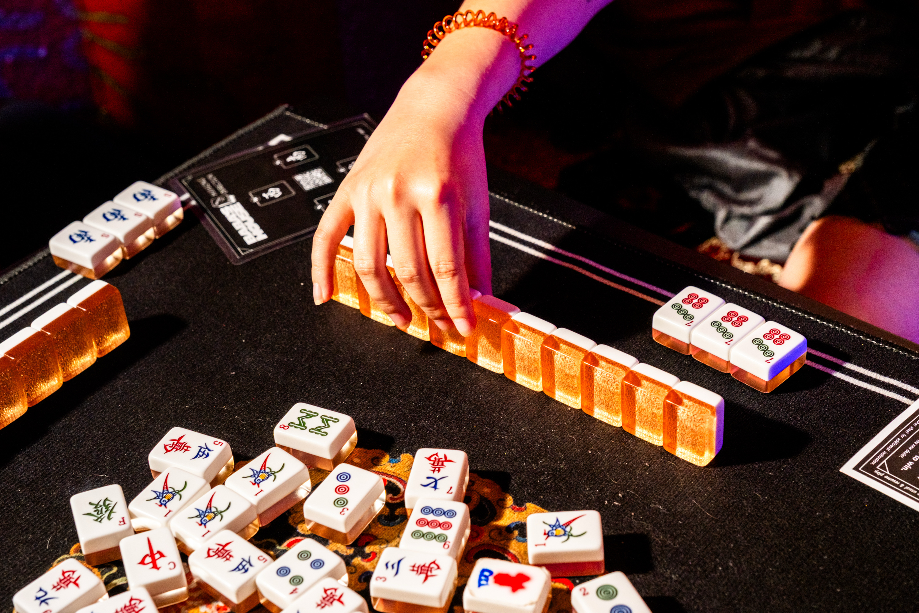 A person with a beaded bracelet adjusts a row of vibrant mahjong tiles on a dark table, surrounded by scattered tiles, under warm lighting.
