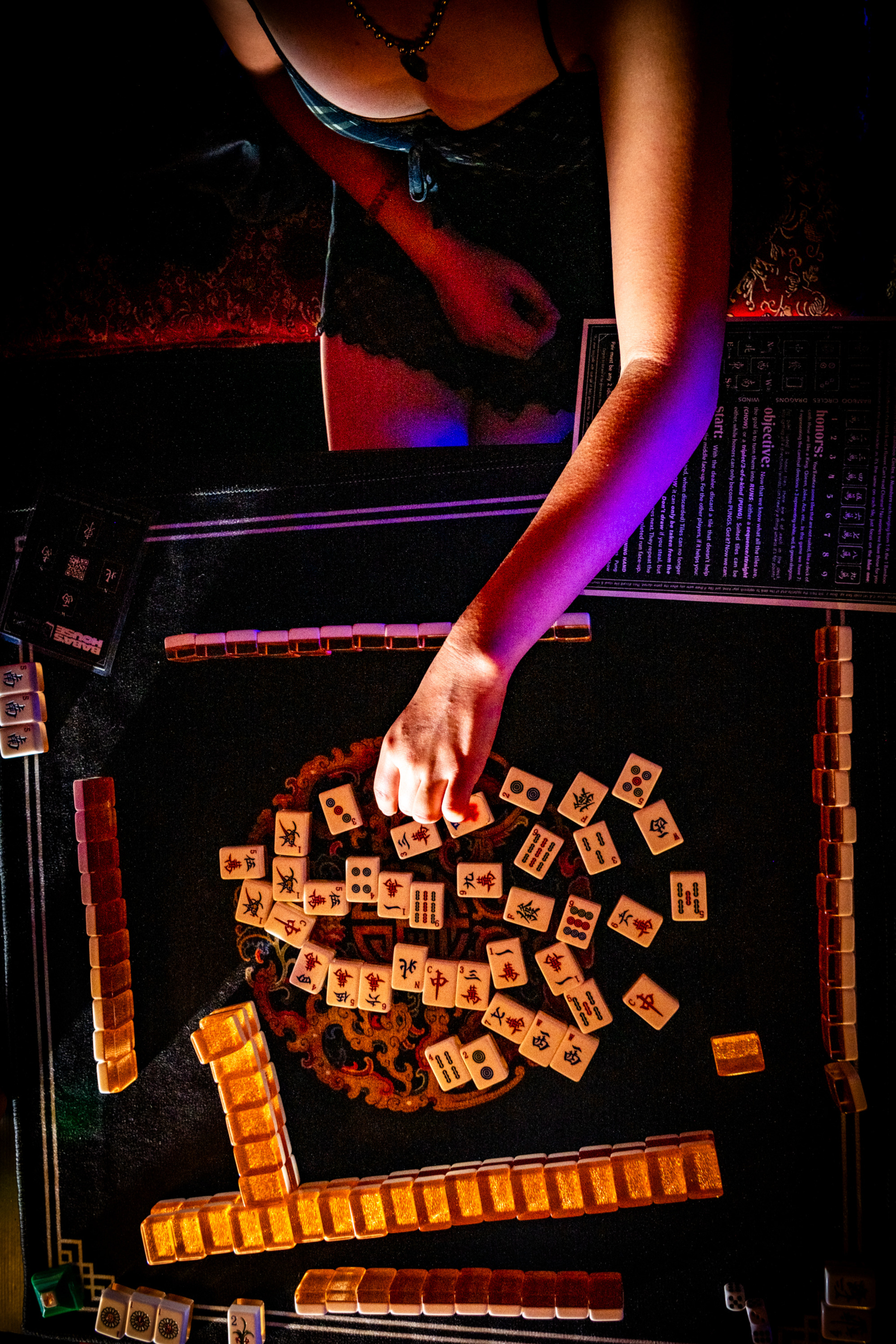 A person is playing Mahjong at a table with scattered tiles in an intricate pattern. Their hand hovers over the tiles, and game pieces line the perimeter.