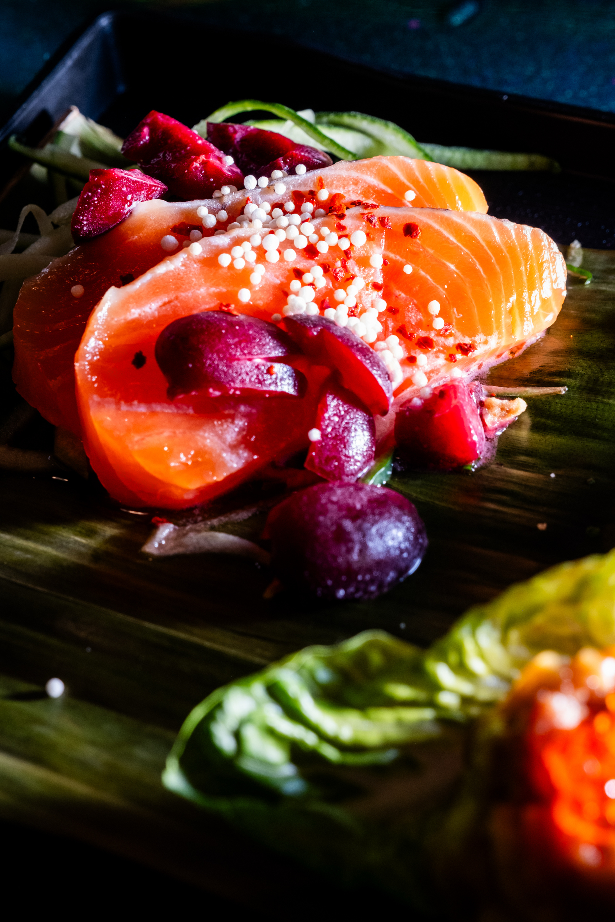 Slices of fresh salmon are artfully arranged with vibrant red fruits and sprinkled with small white beads, presented on a leaf-lined platter.