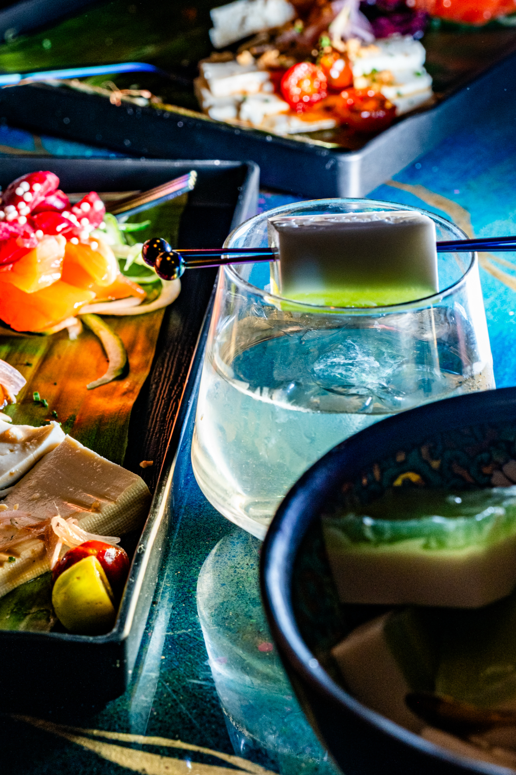 A vibrant spread features colorful sliced vegetables, a glass with a square dessert atop, and several artistically arranged dishes on a dark table.
