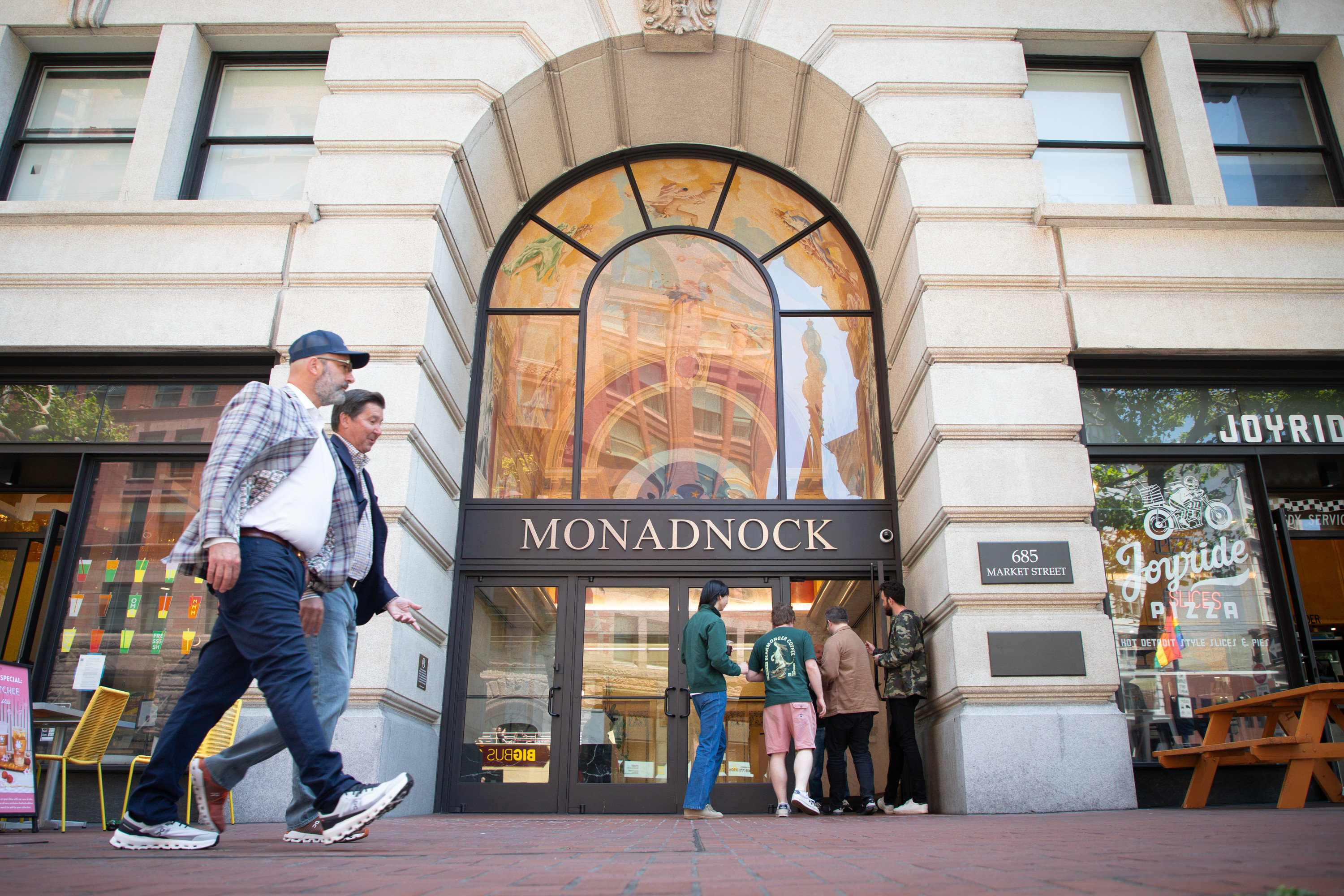 A group of people stands outside the Monadnock building entrance at 685 Market Street. Some are walking past and others gather near the door, with a pizza shop nearby.