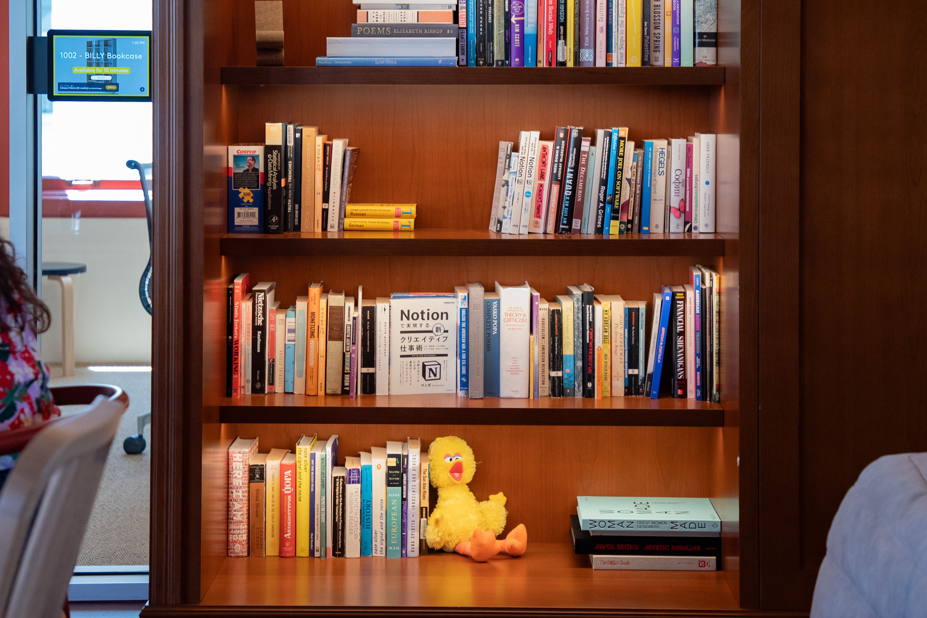 A wooden bookshelf with multiple shelves filled with books. A yellow plush toy, resembling a bird, is on the middle shelf. A digital screen is visible on the left.