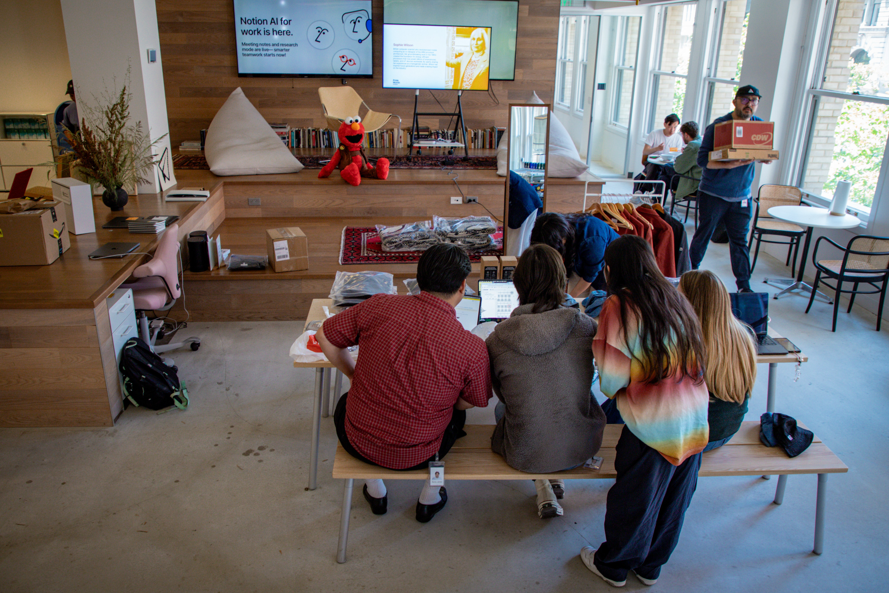 A group of people sits around a table with laptops in a bright, modern office. A plush toy of Elmo is on a wooden bench by the wall, below two screens.