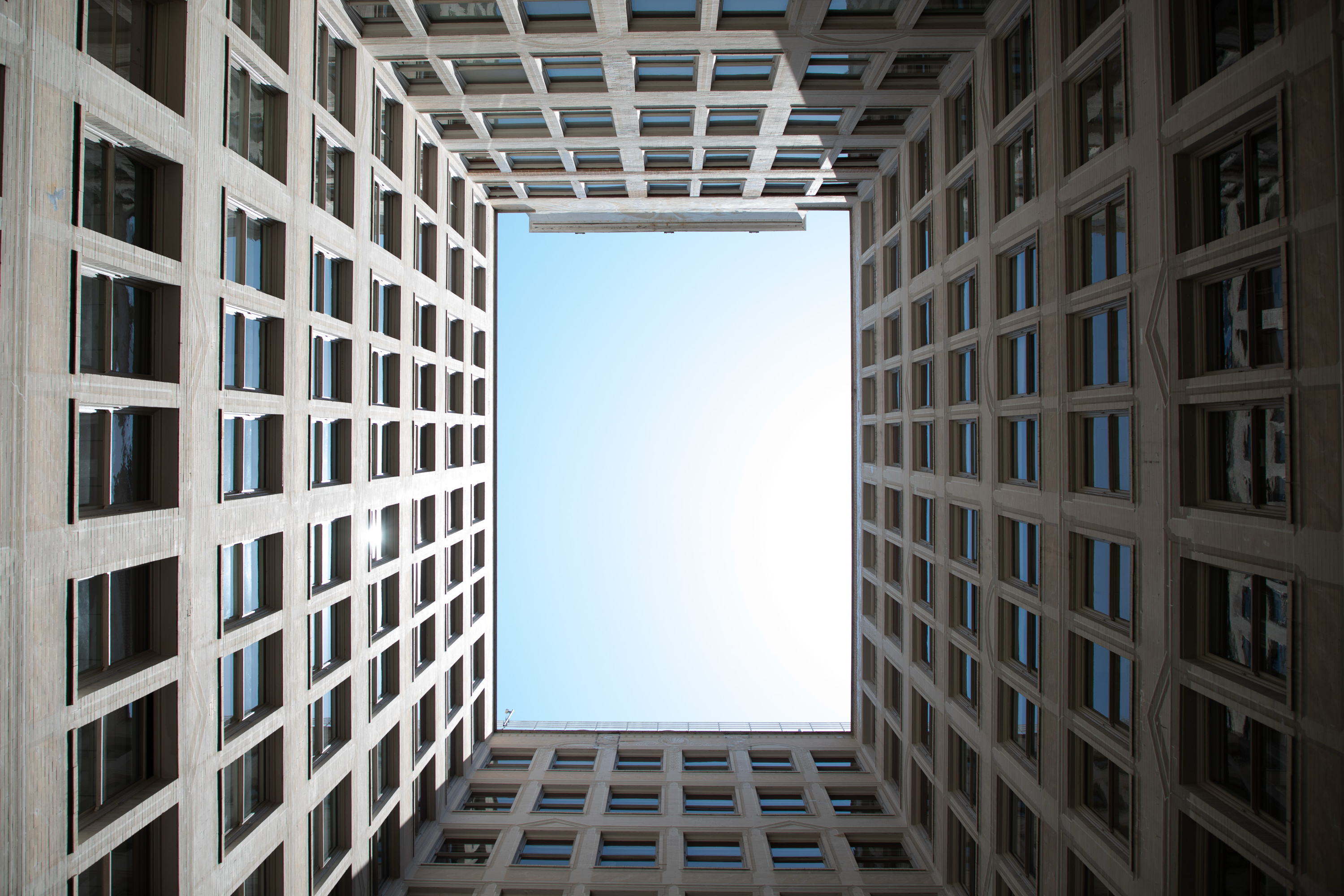 The image shows a view looking up at the sky from within a square courtyard surrounded by high-rise buildings with repeating window patterns.