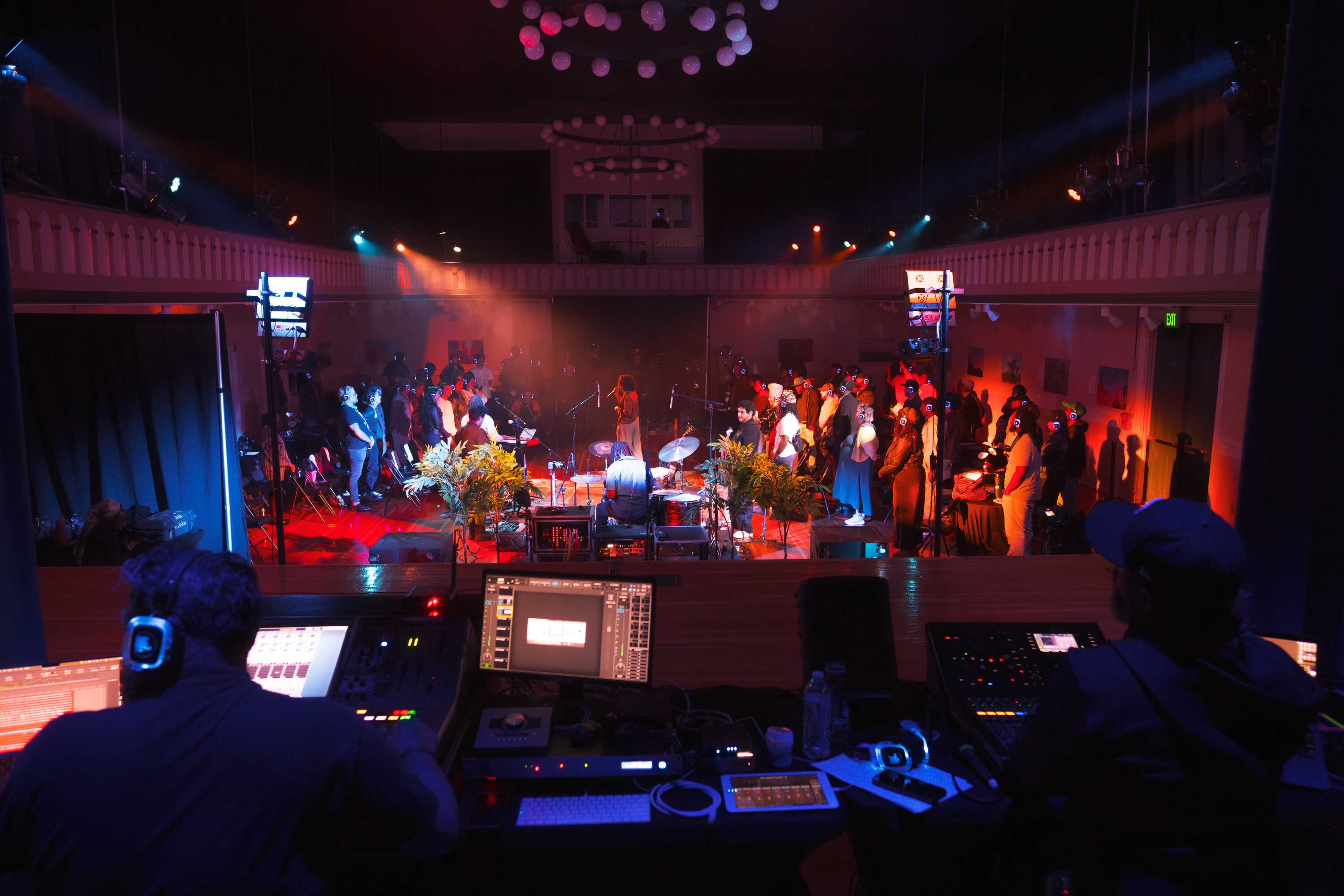 A concert setting featuring a band playing on stage, surrounded by an audience wearing headphones. The scene is lit with colorful stage lights.