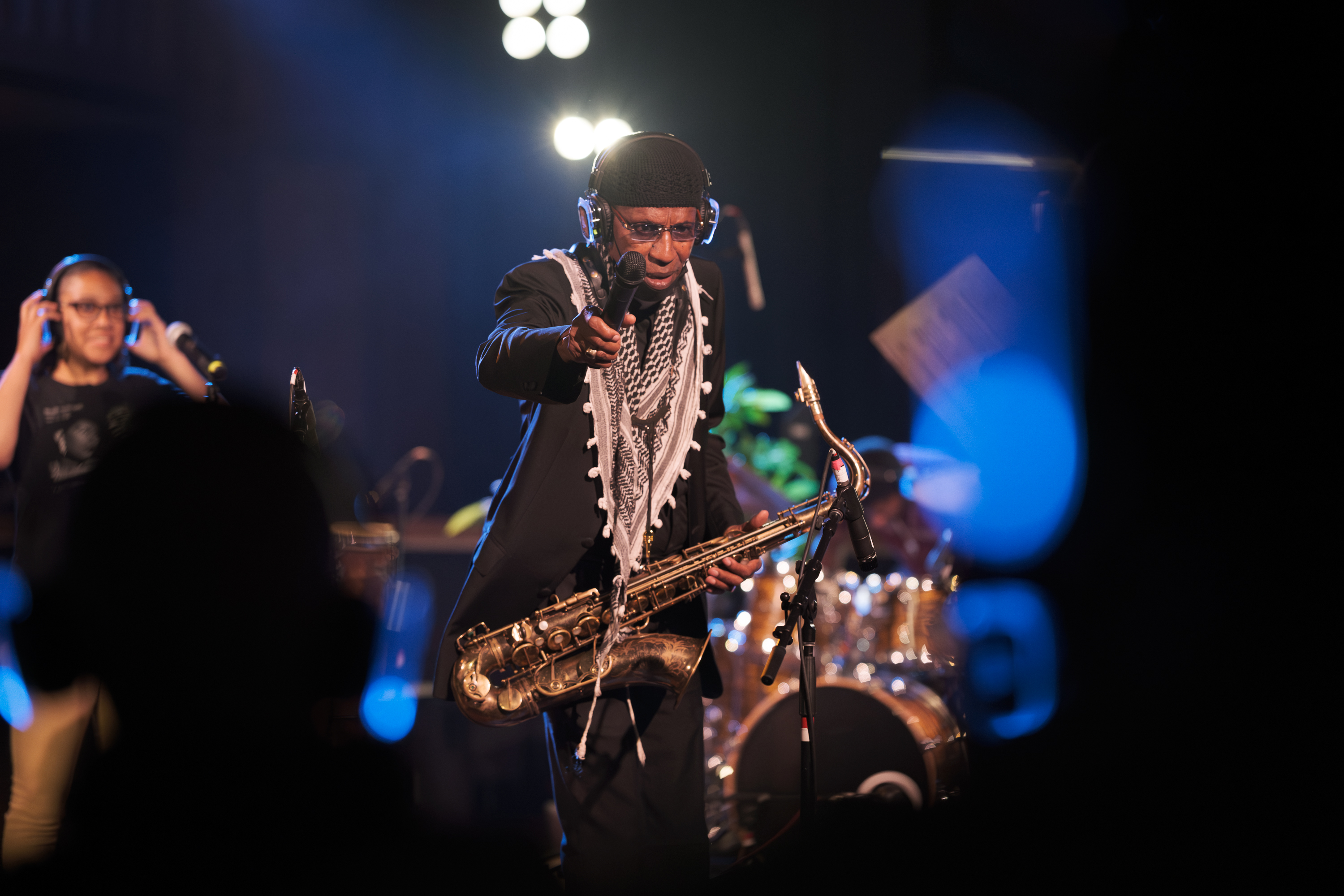 A musician on stage, wearing a patterned scarf and headphones, points a microphone towards the audience while holding a saxophone. Bright stage lights illuminate the scene.