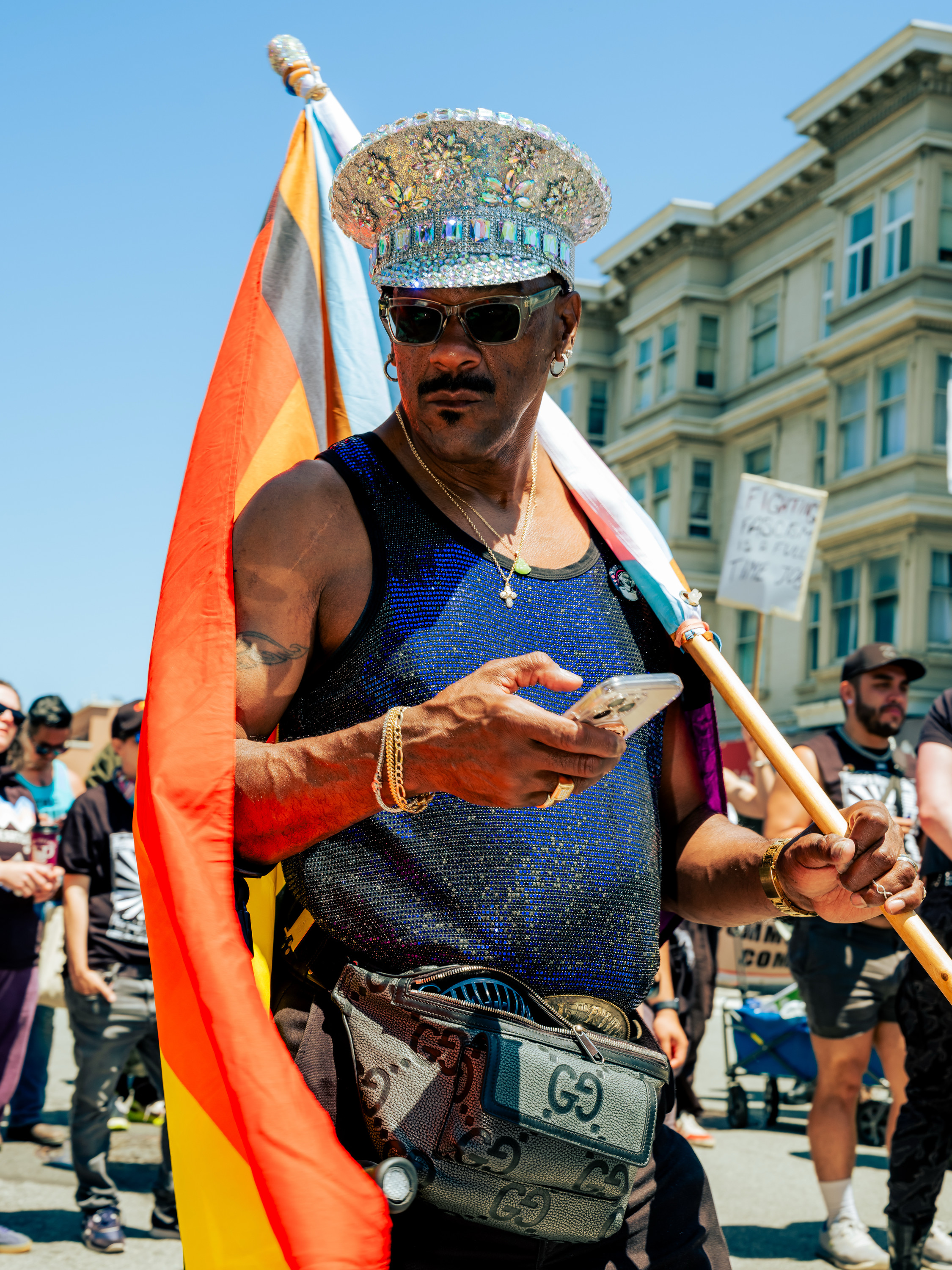A person wearing a sparkly hat and sunglasses holds a phone and flag. They stand in front of a building, with a colorful crowd and sign in the background.