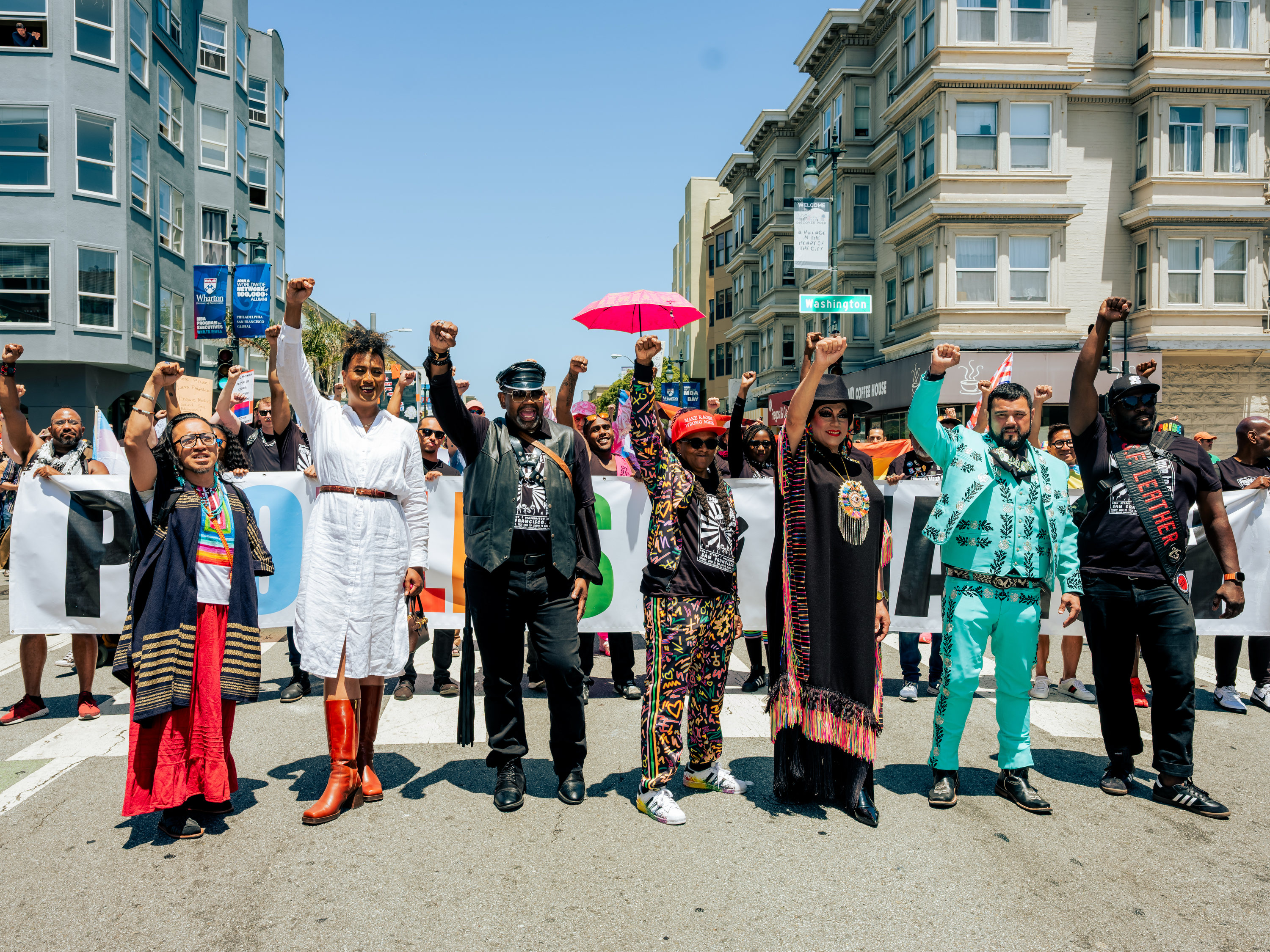 A diverse group of people stands on a street, arms raised in unity, holding a banner. The background shows city buildings and the clear blue sky above.
