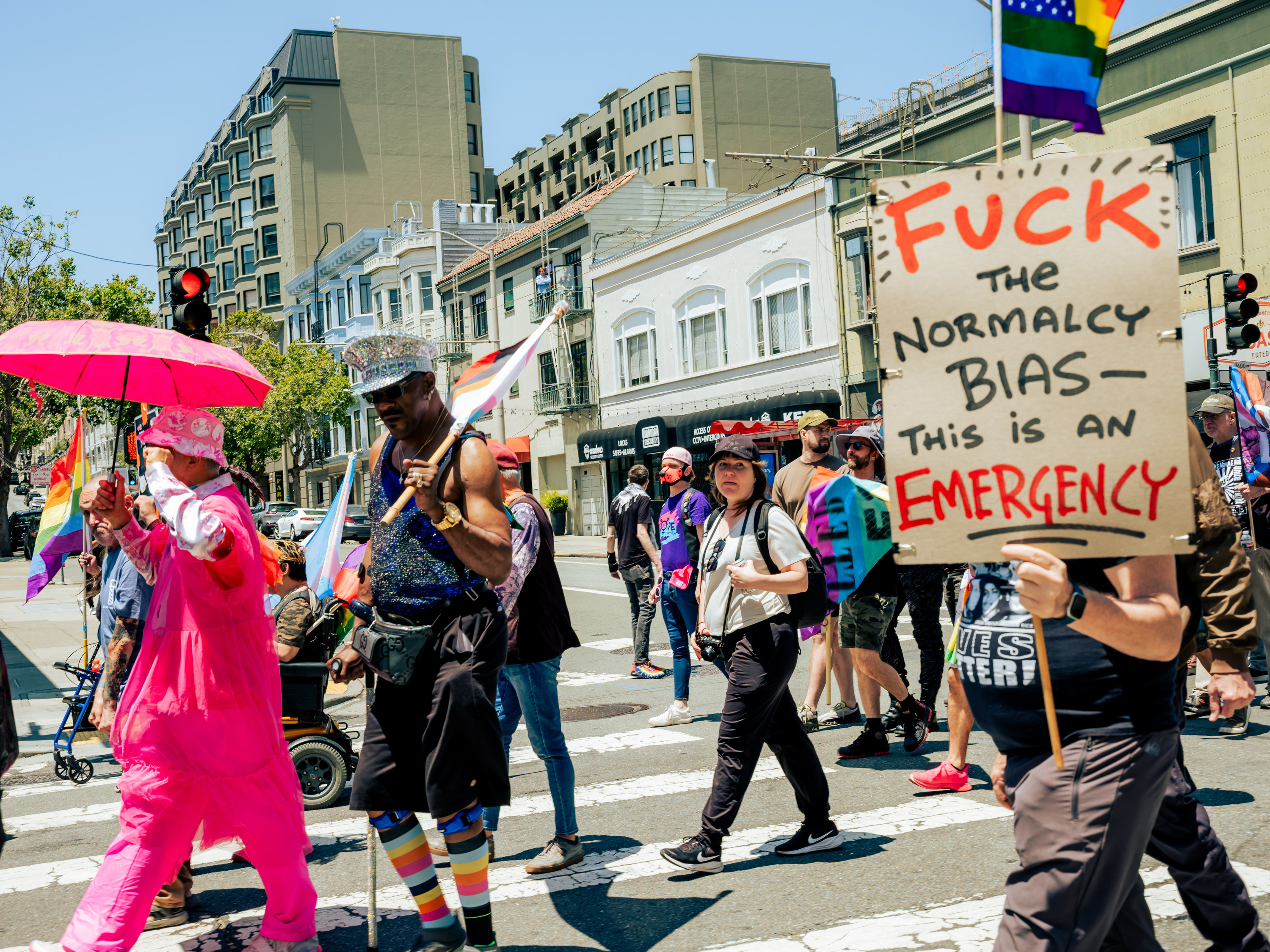 The image shows a lively parade with people in colorful outfits. One person holds a sign reading, &quot;F*** the Normalcy Bias—This is an Emergency.&quot;