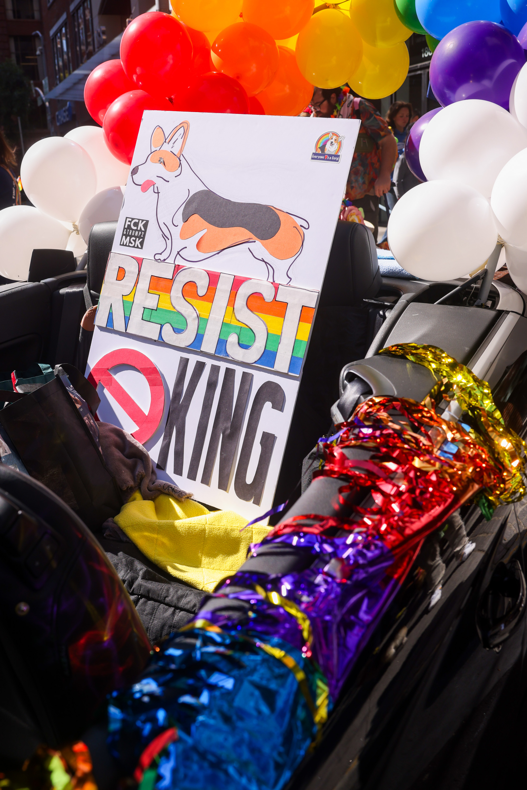 A colorful float features a corgi illustration, rainbow &quot;RESIST&quot; text, and rainbow streamers, surrounded by red, yellow, and white balloons.