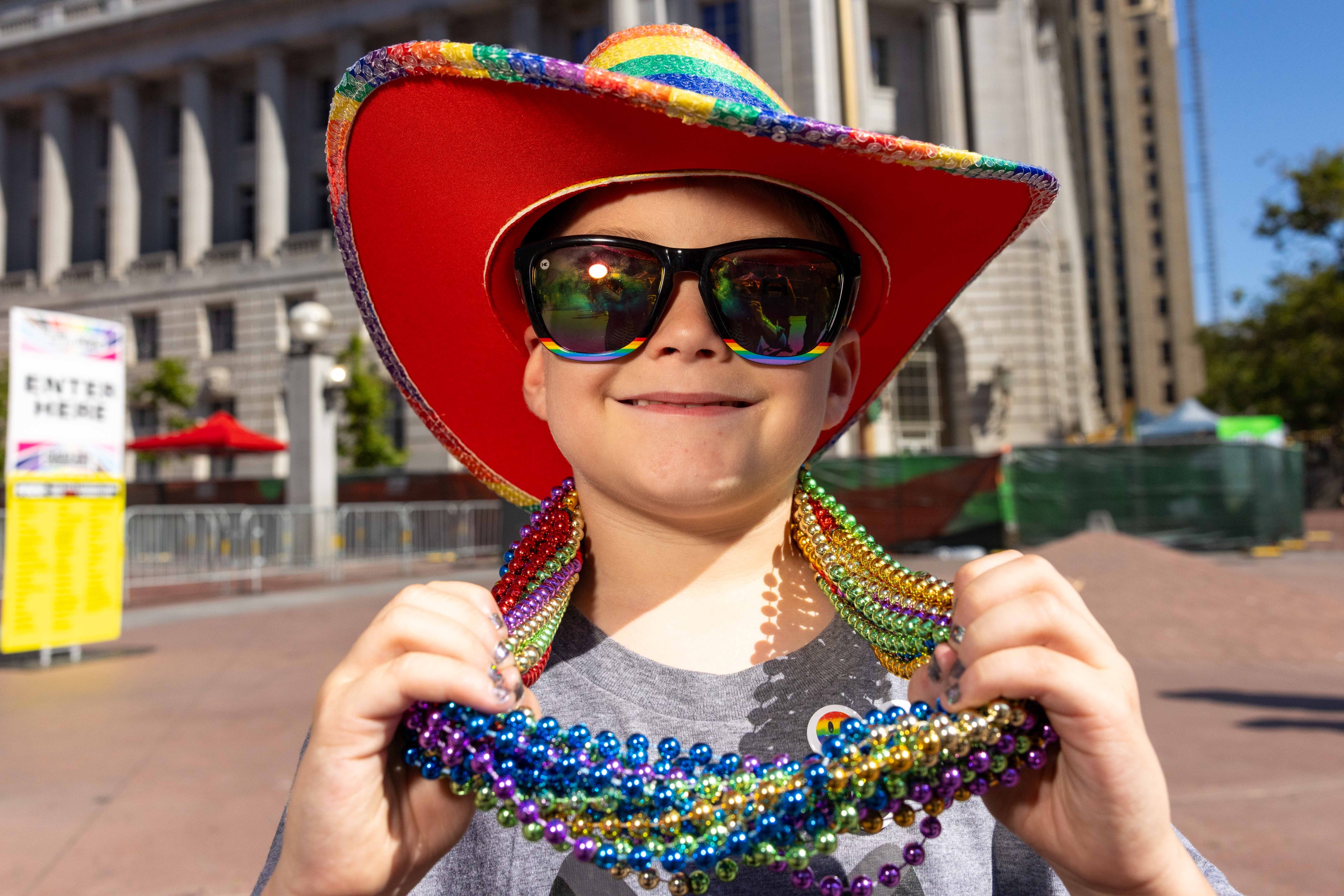 A child wearing a rainbow cowboy hat and sunglasses smiles while holding colorful beaded necklaces. The background shows a building and a sign that says &quot;ENTER HERE&quot;.