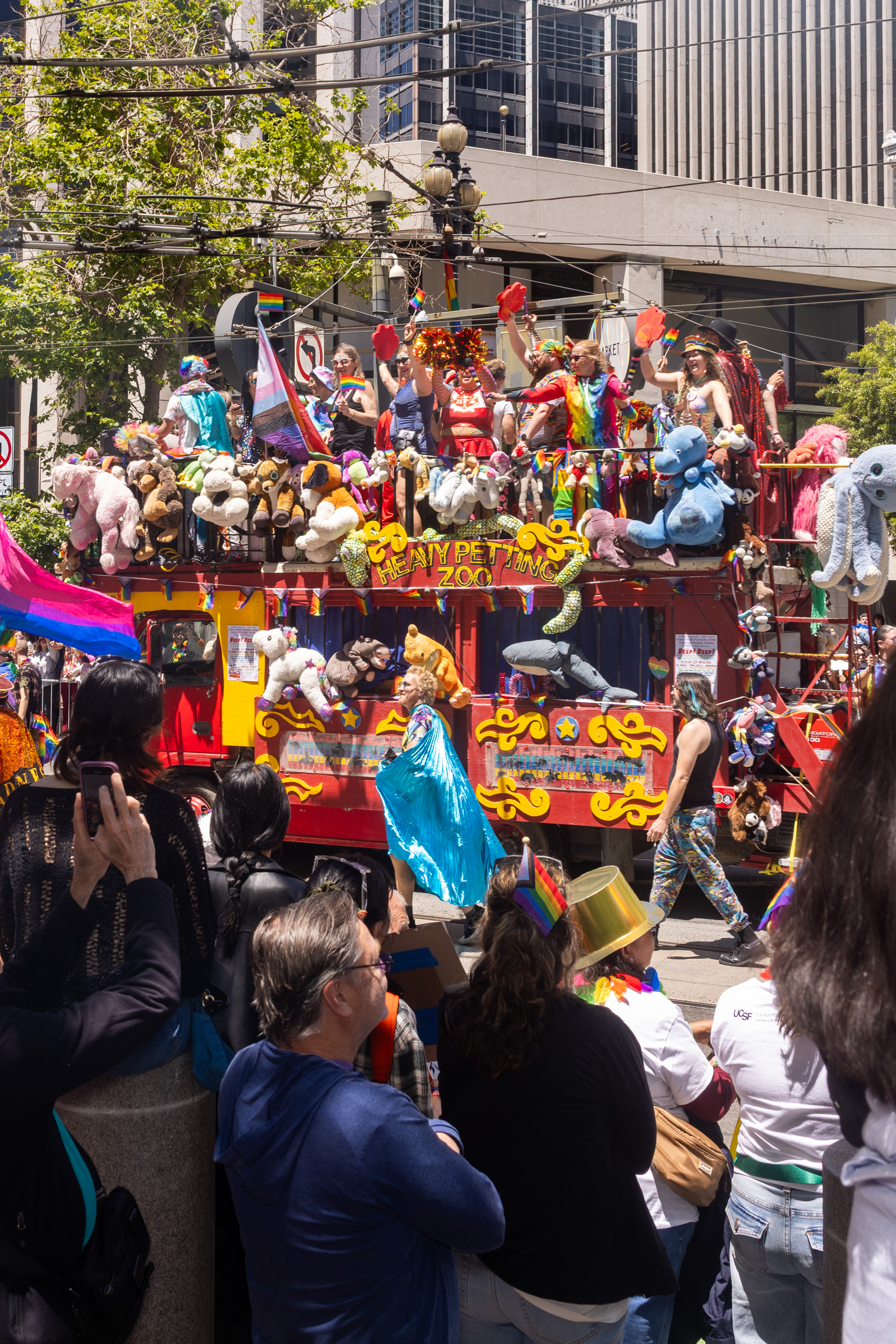 A colorful parade float covered in stuffed animals and rainbow decorations passes by a crowd. People on the float are cheering and waving flags.