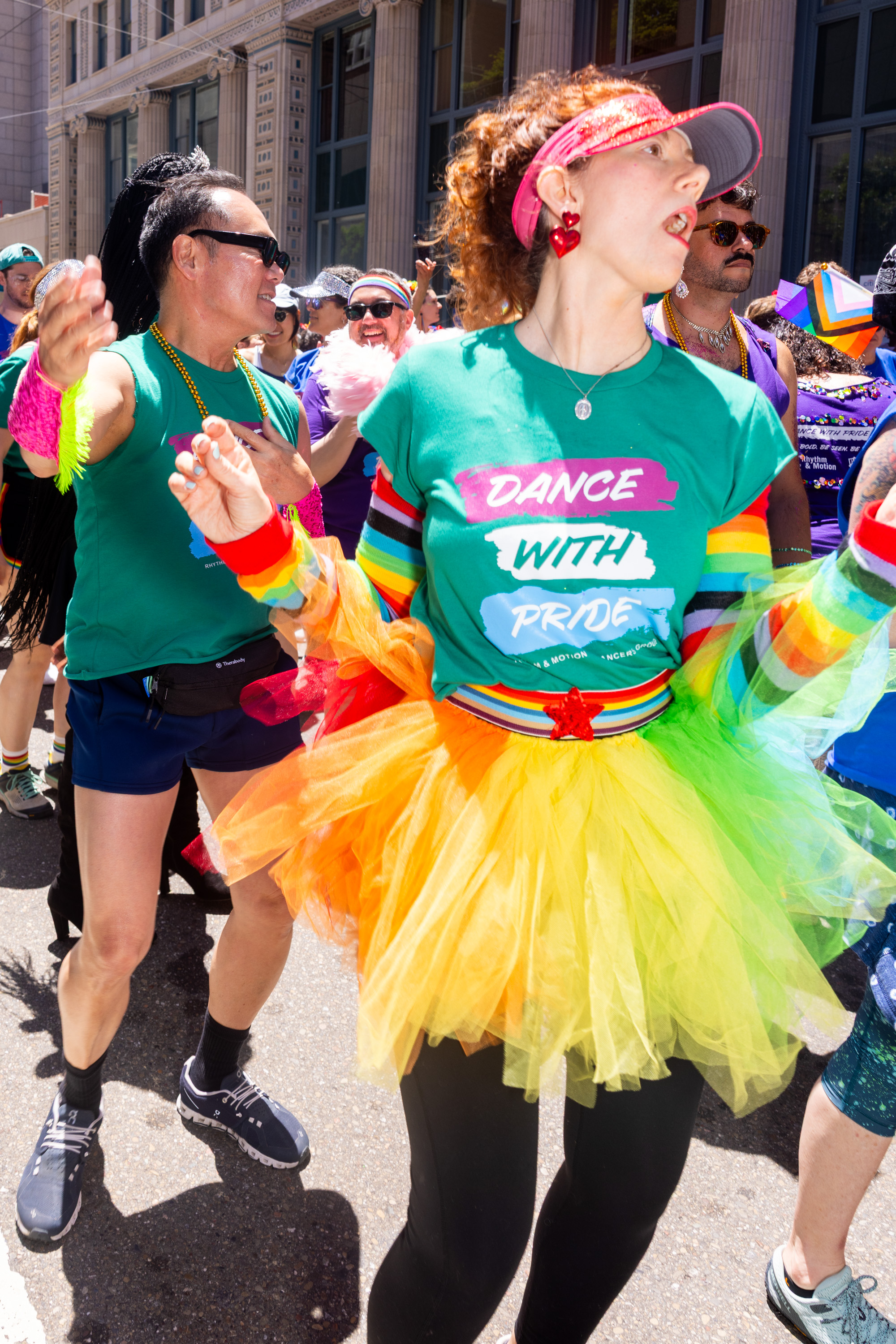 People in colorful outfits and accessories joyfully dance at a parade. A woman wears a shirt reading &quot;Dance with Pride&quot; and a vibrant tutu.