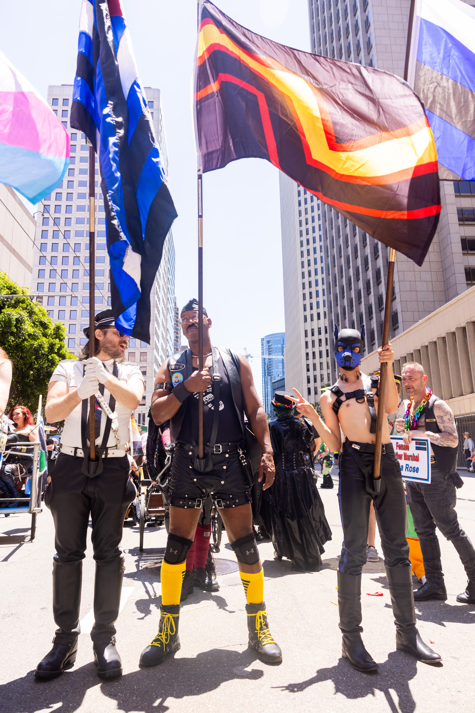 People in diverse, colorful outfits hold flags at an outdoor event, standing tall among city buildings and concrete structures.