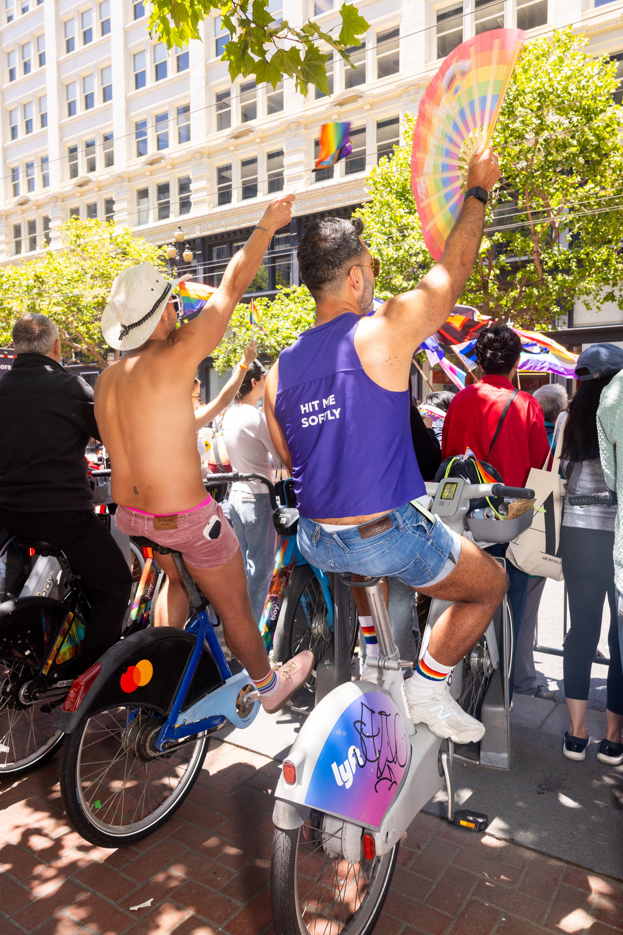 People on bikes celebrate in a city street, waving rainbow flags. One person has a rainbow fan and wears a &quot;Hit Me Soflty&quot; shirt. Trees and a building are visible.