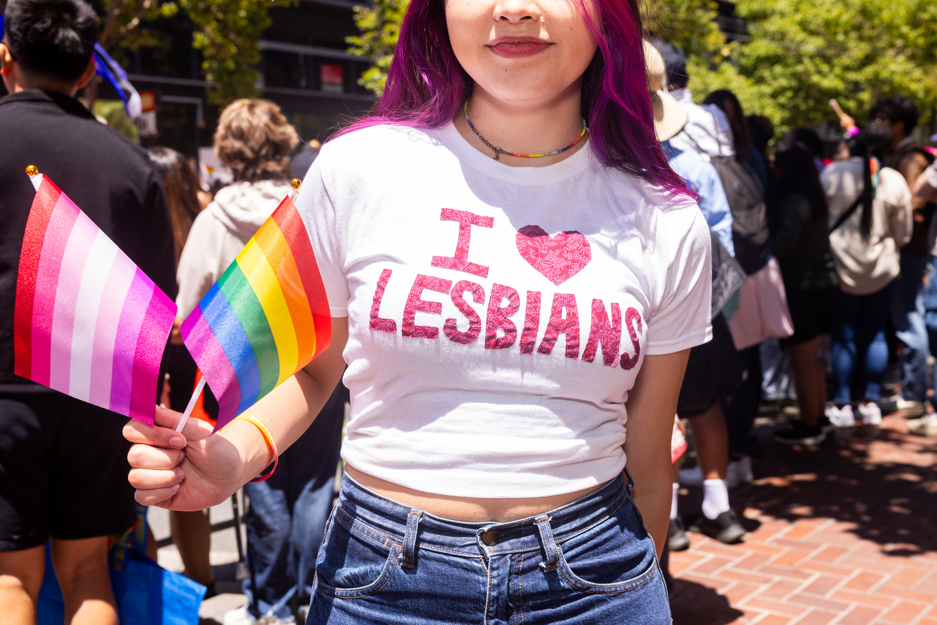 A person with purple hair wears a white shirt saying &quot;I ♥ LESBIANS&quot; and holds lesbian and rainbow flags, standing in a crowded outdoor setting.