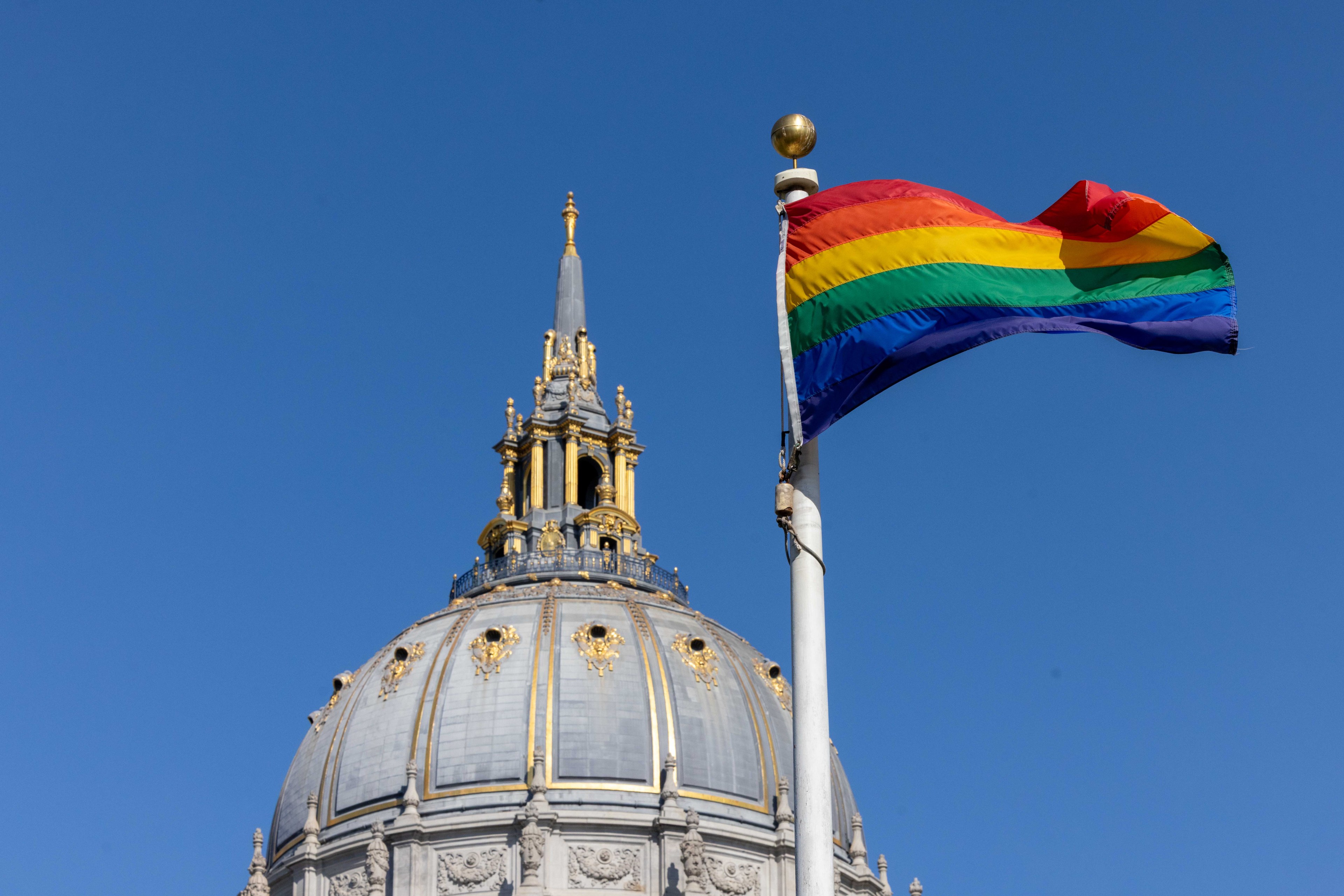 A domed building with ornate gold details stands against a clear blue sky, beside a pole with a rainbow pride flag waving.