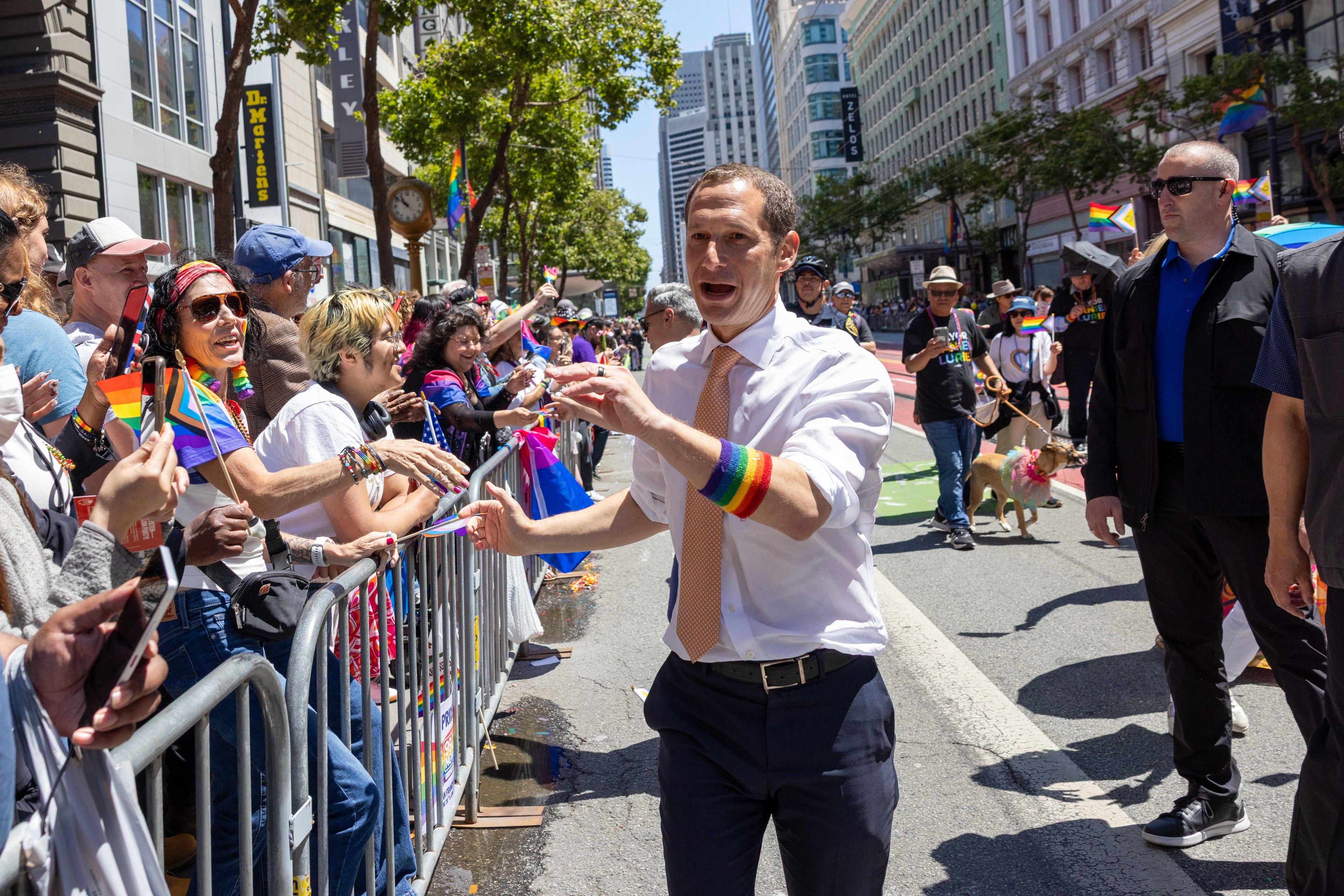 A person wearing a white shirt and rainbow armband interacts with a joyful crowd behind a barricade at a vibrant outdoor event on a city street.