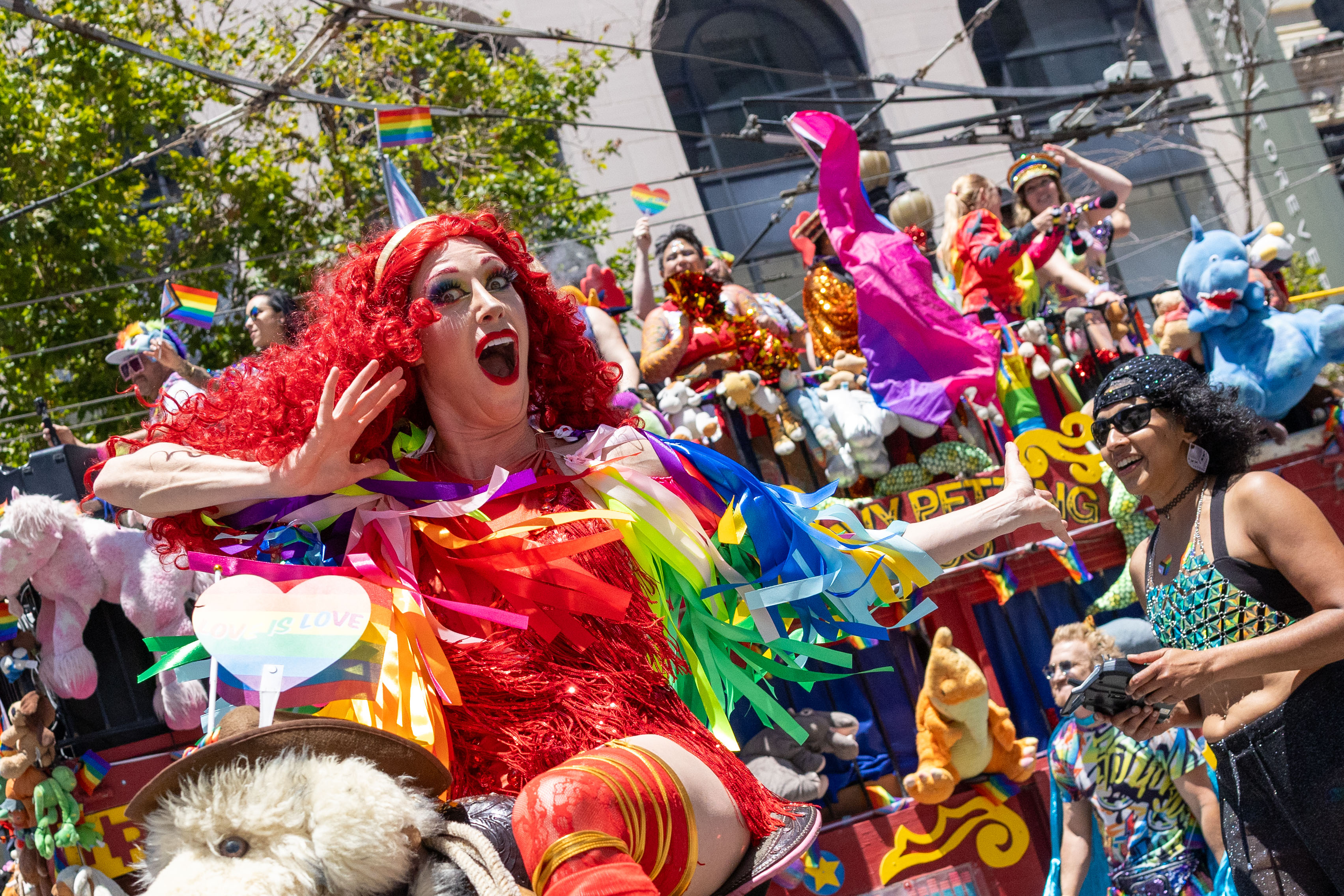 A person with bright red hair, colorful ribbons, and a vibrant outfit is joyfully posing in a parade. They're surrounded by stuffed animals and pride flags.