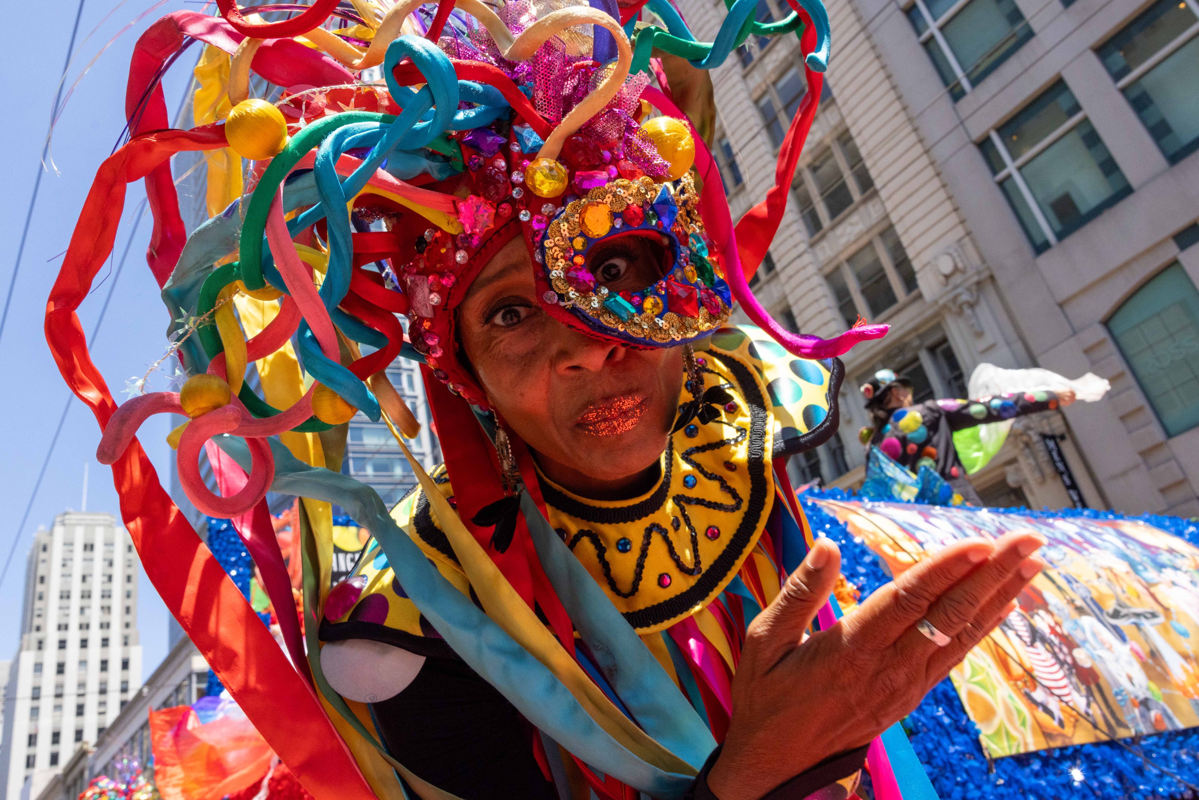 A person in a colorful, elaborate costume with ribbons and a decorated mask is performing in a vibrant street parade in a cityscape setting.