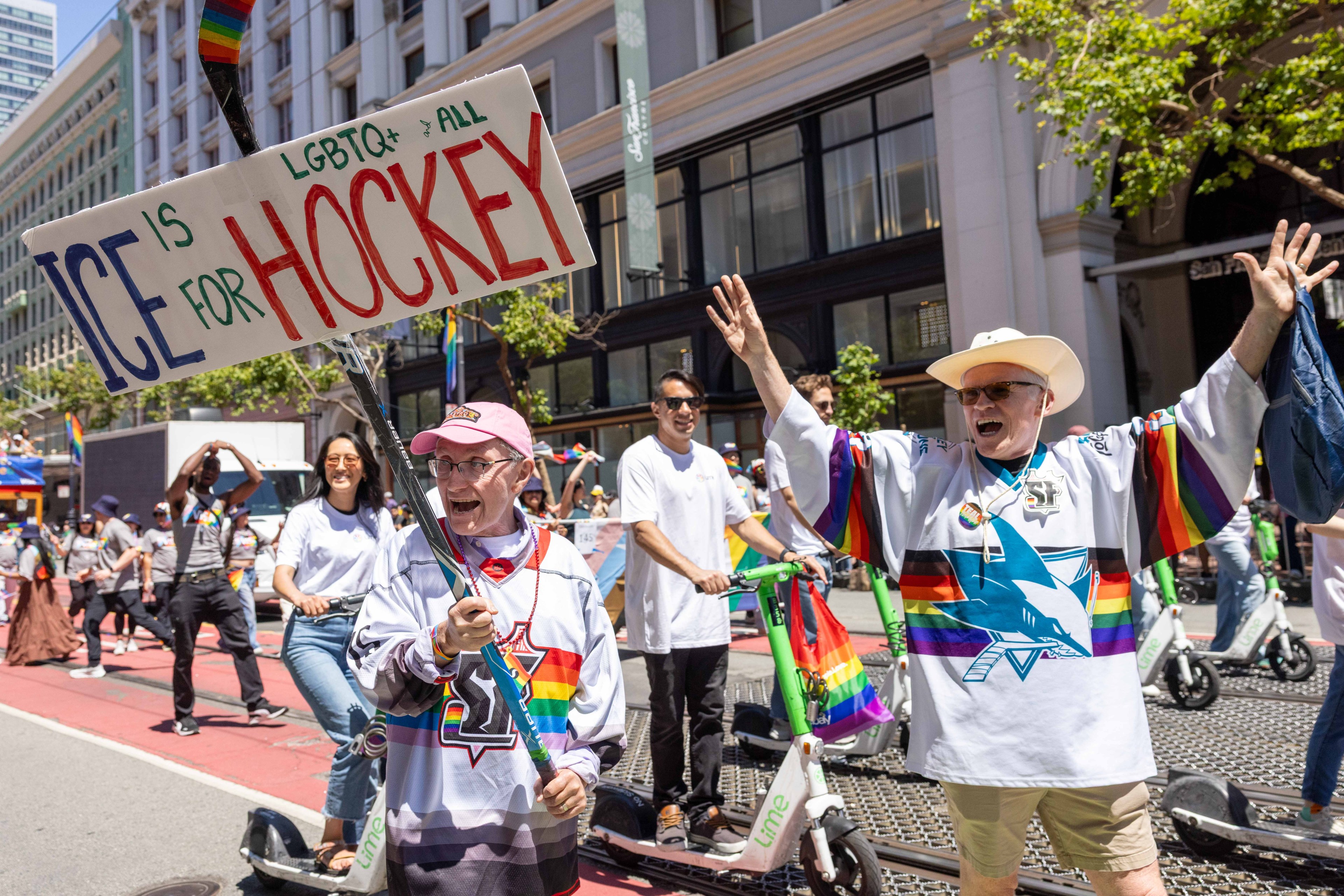 A lively parade scene with people in colorful attire, waving a sign saying, &quot;Ice is for Hockey.&quot; They are smiling and standing beside scooters on a sunny street.