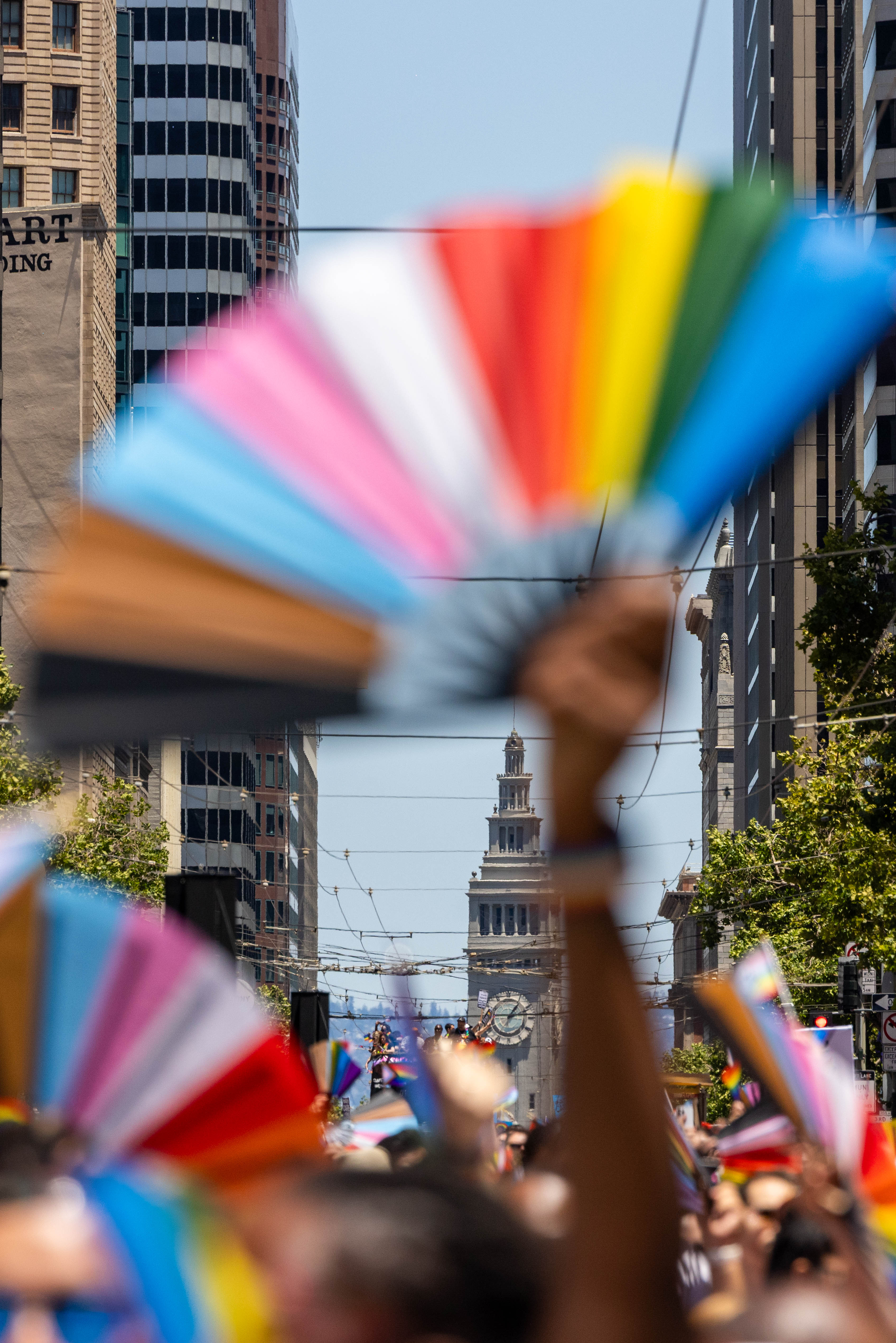 The image shows a vibrant city street scene with people holding colorful, rainbow-themed fans. The background features a prominent tower amidst tall buildings.