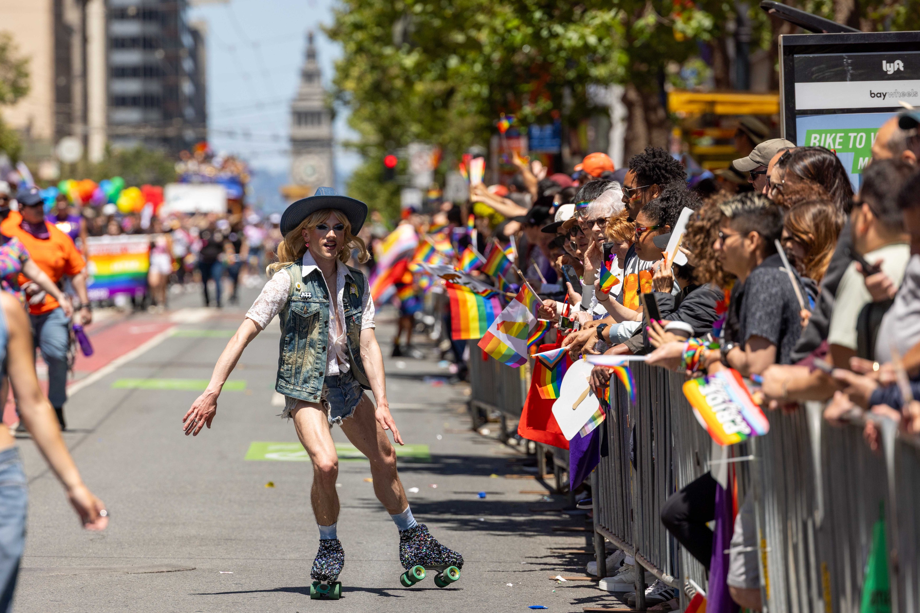 A person on roller skates in a denim vest and shorts skates along a street during a vibrant parade. Crowd members wave rainbow flags, creating a festive atmosphere.