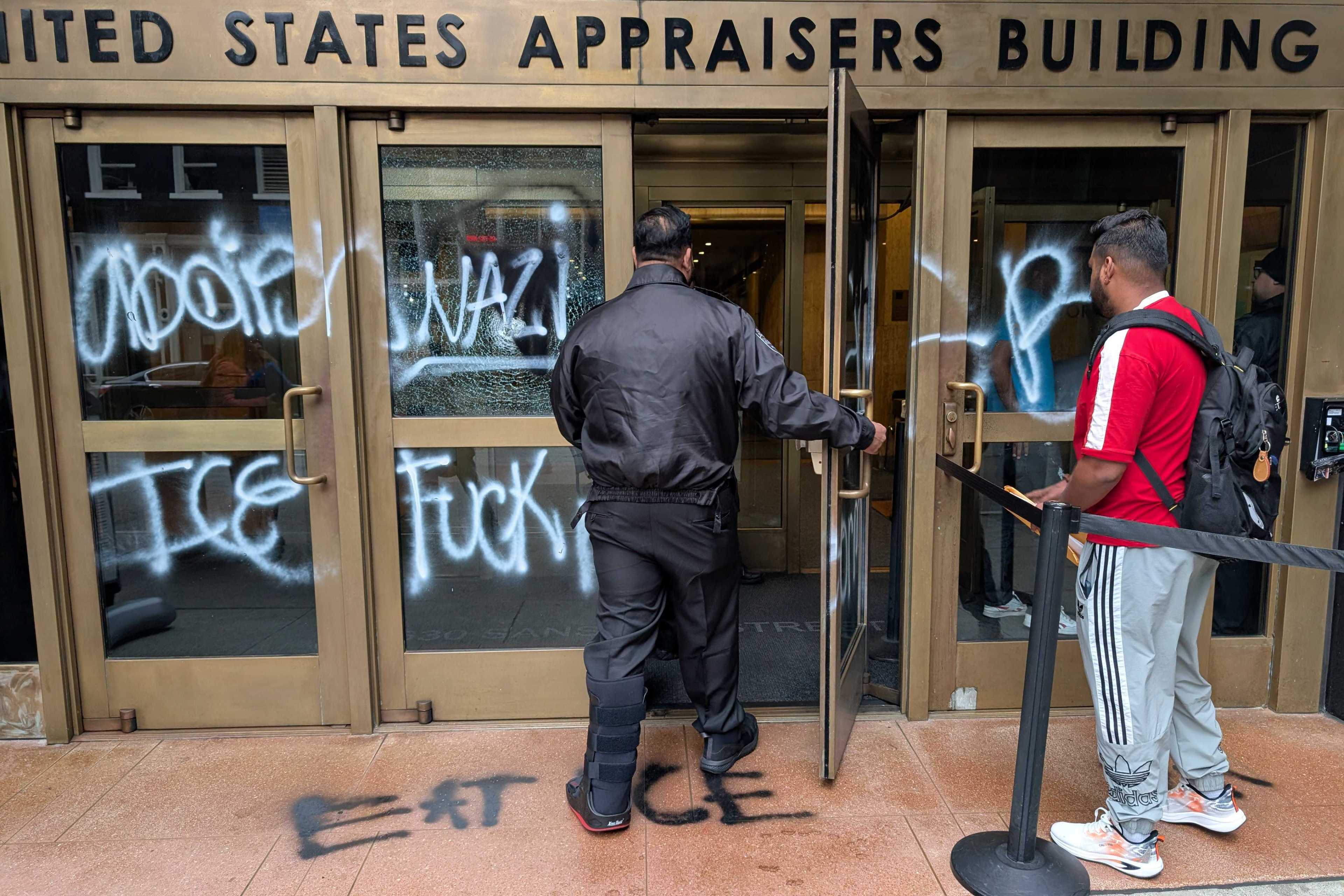 The image shows the entrance of the United States Appraisers Building with graffiti on the glass doors. Two people are entering, one wearing a red shirt and backpack.