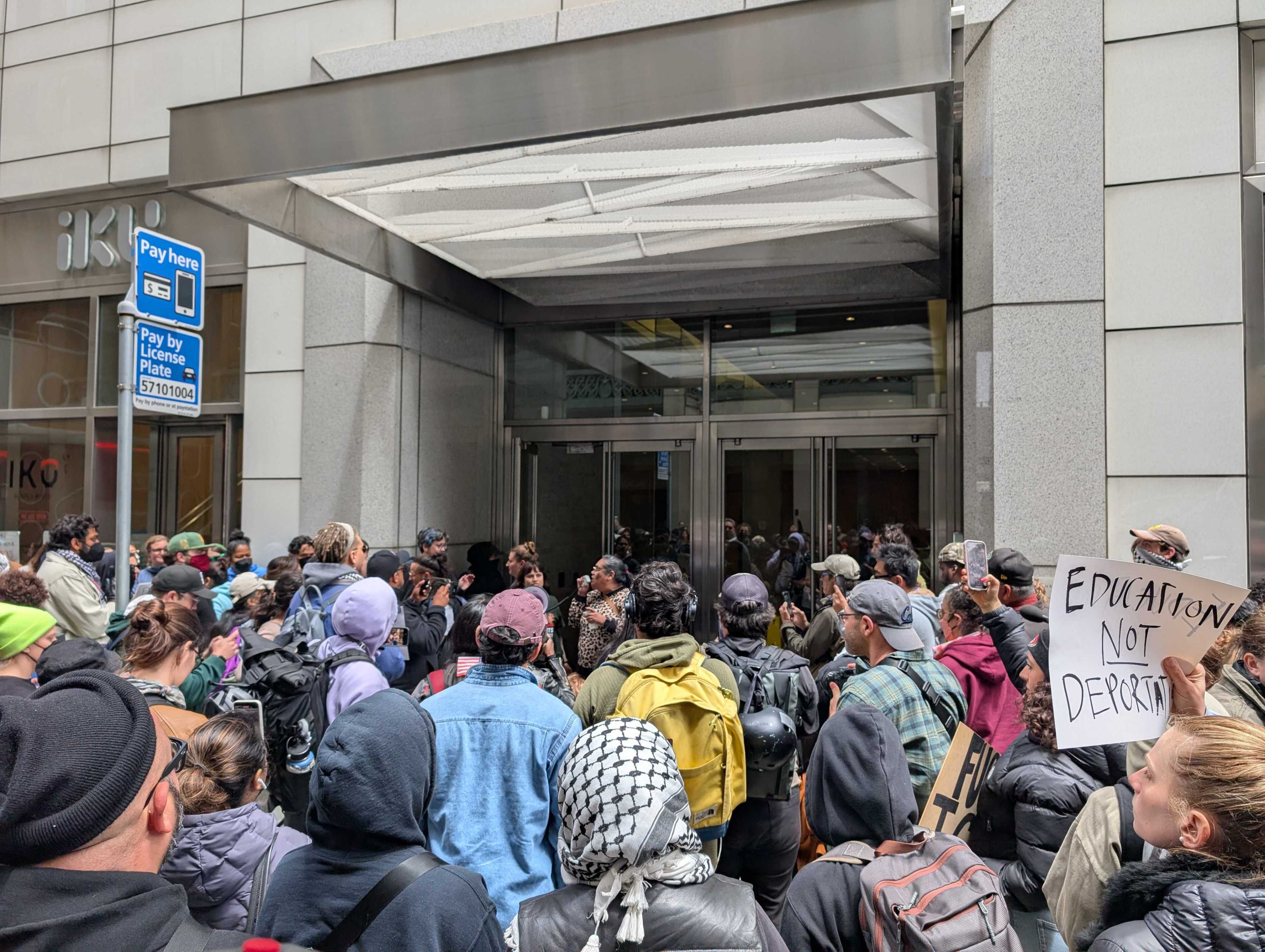 A large group of people gathers outside a building entrance, holding signs with messages like &quot;Education Not Deportation,&quot; suggesting a protest or demonstration.