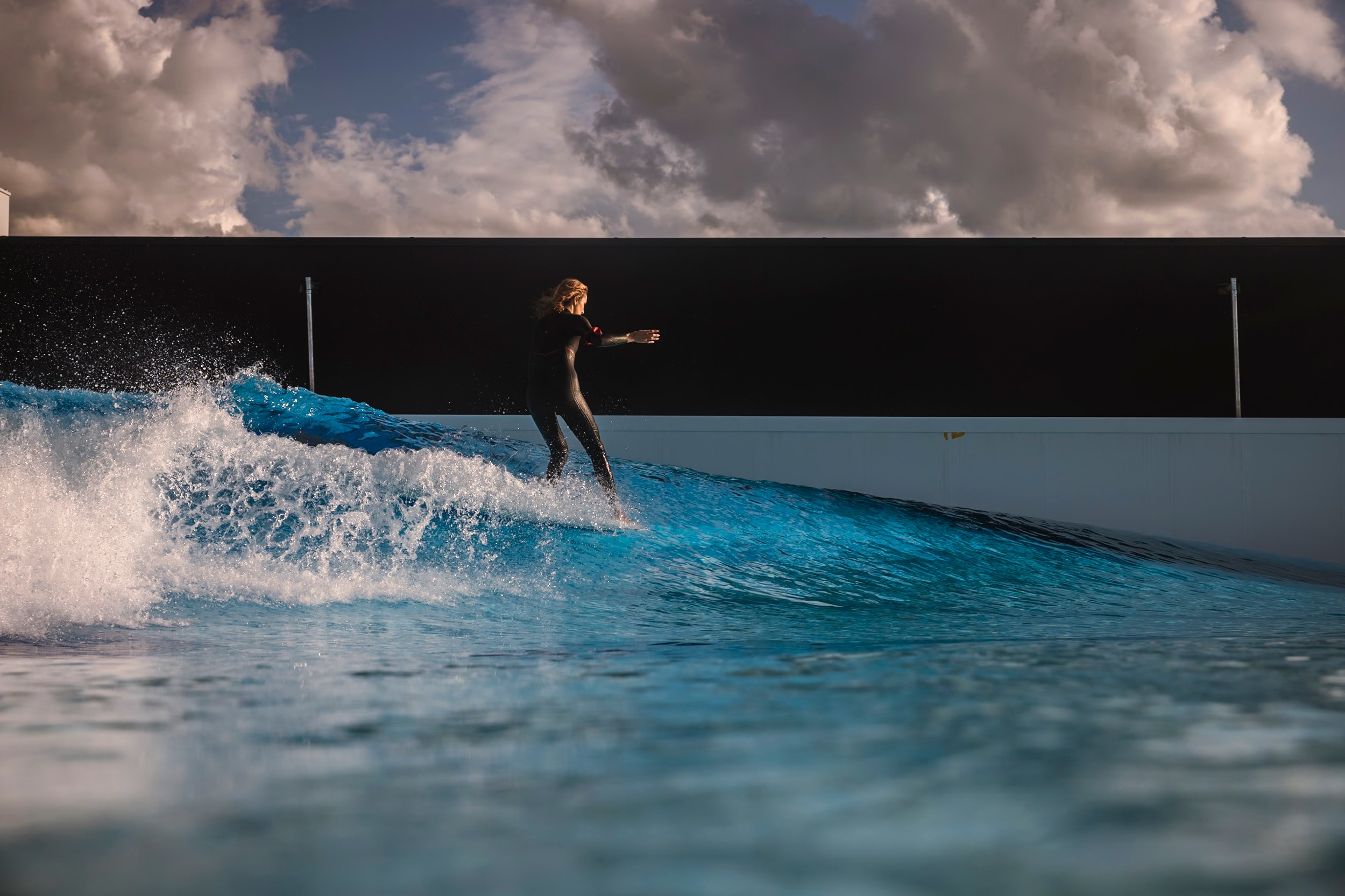 A person in a wetsuit surfs on a bright blue artificial wave under a dramatic cloudy sky, against a modern structure's backdrop.