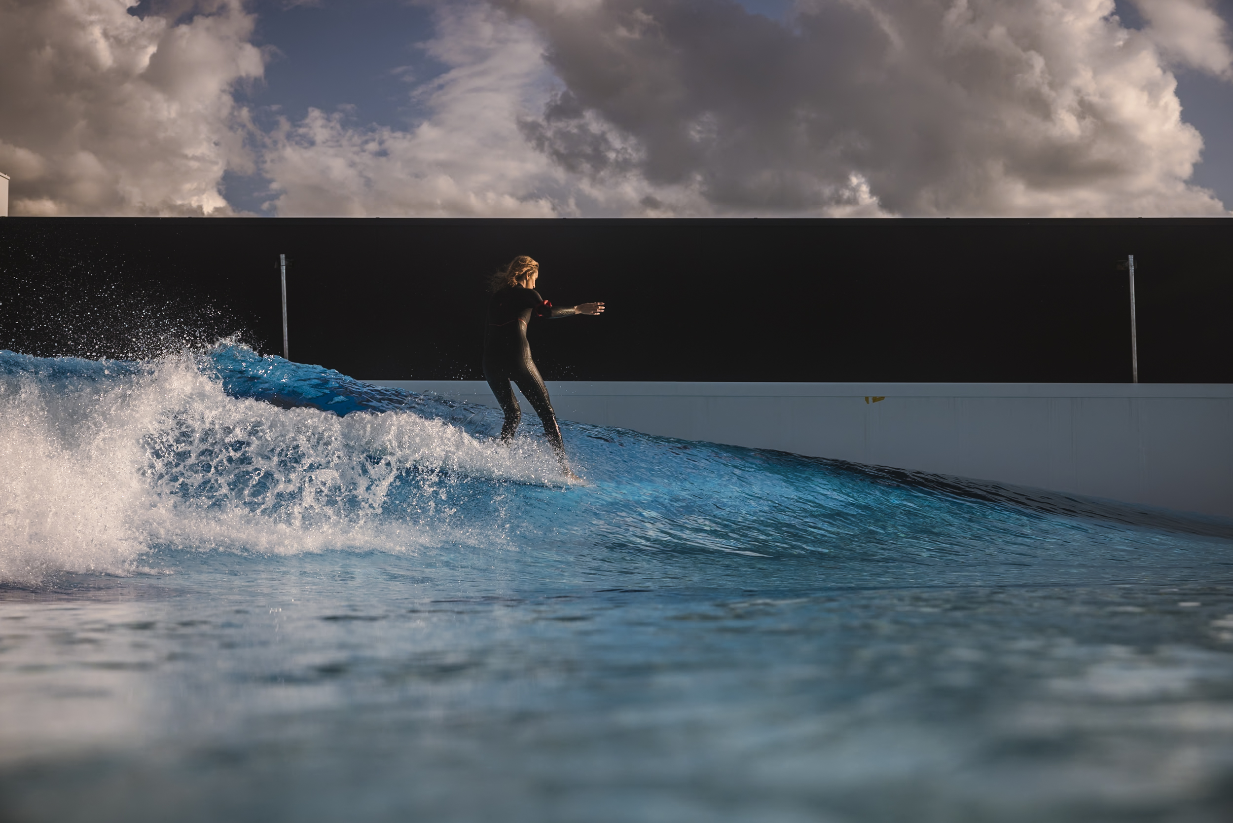 A person in a wetsuit surfs on a bright blue artificial wave under a dramatic cloudy sky, against a modern structure's backdrop.