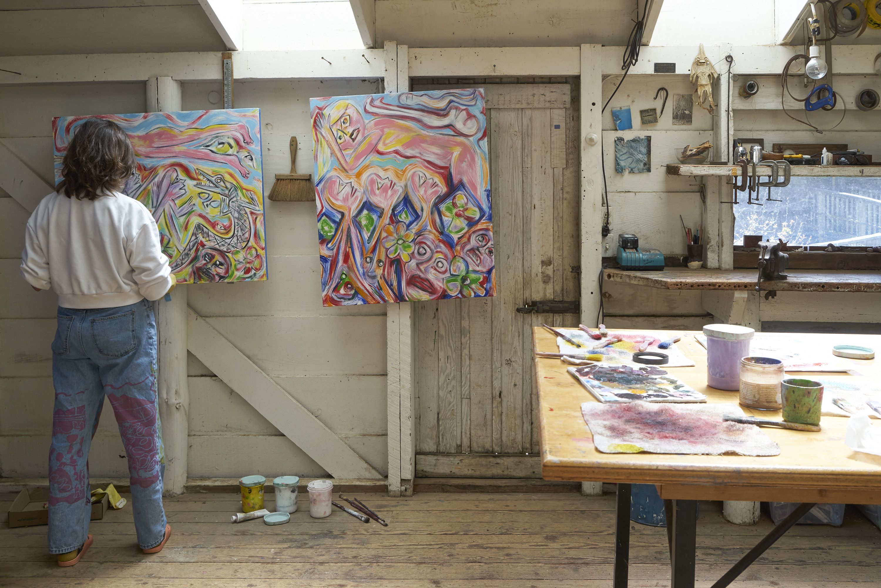 A person stands in an art studio, painting colorful abstract designs on canvases. The room is filled with art supplies and natural light from skylights.