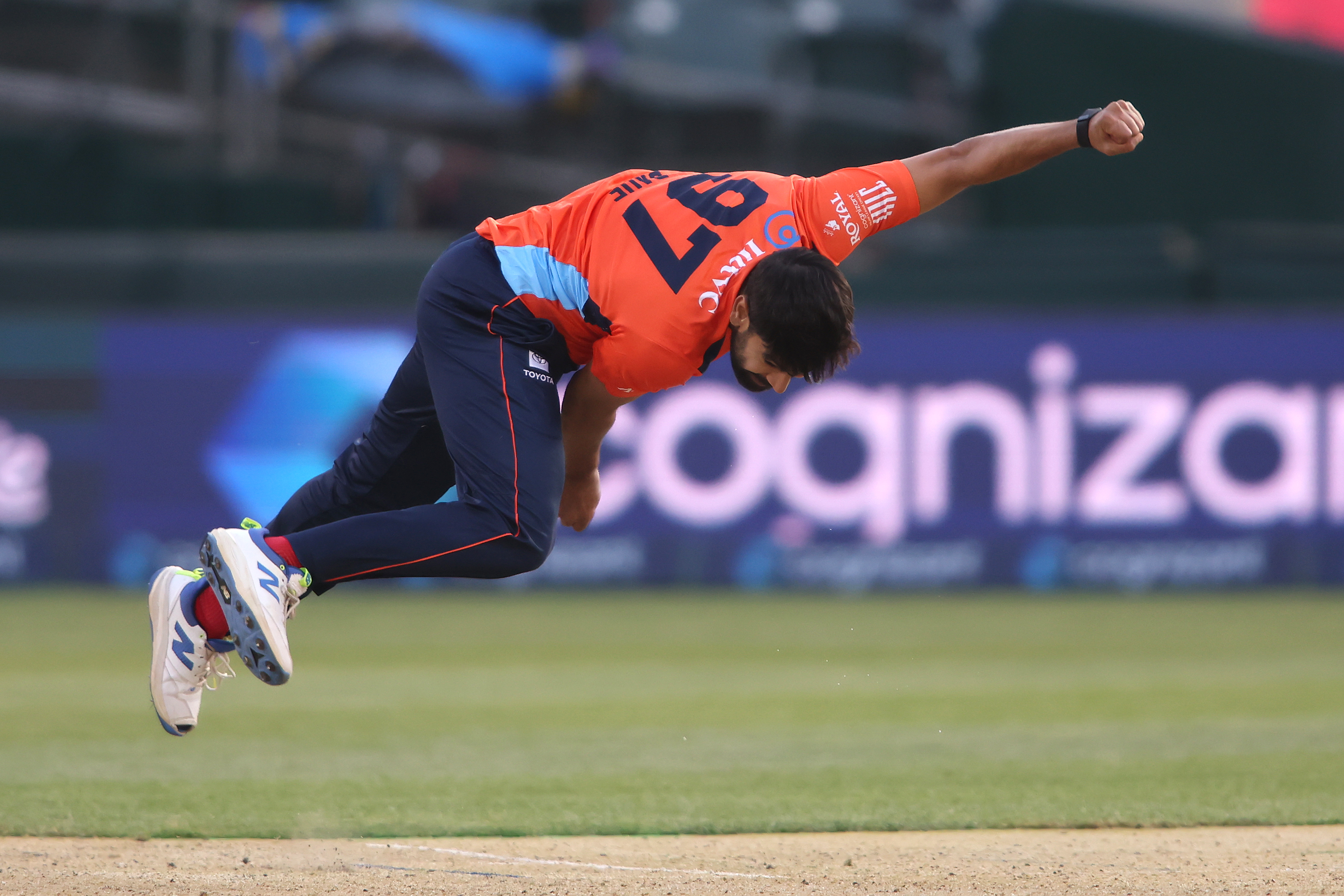 A cricket player in an orange and blue uniform is captured mid-action bowling on the field. He is bent forward, with one foot slightly off the ground.