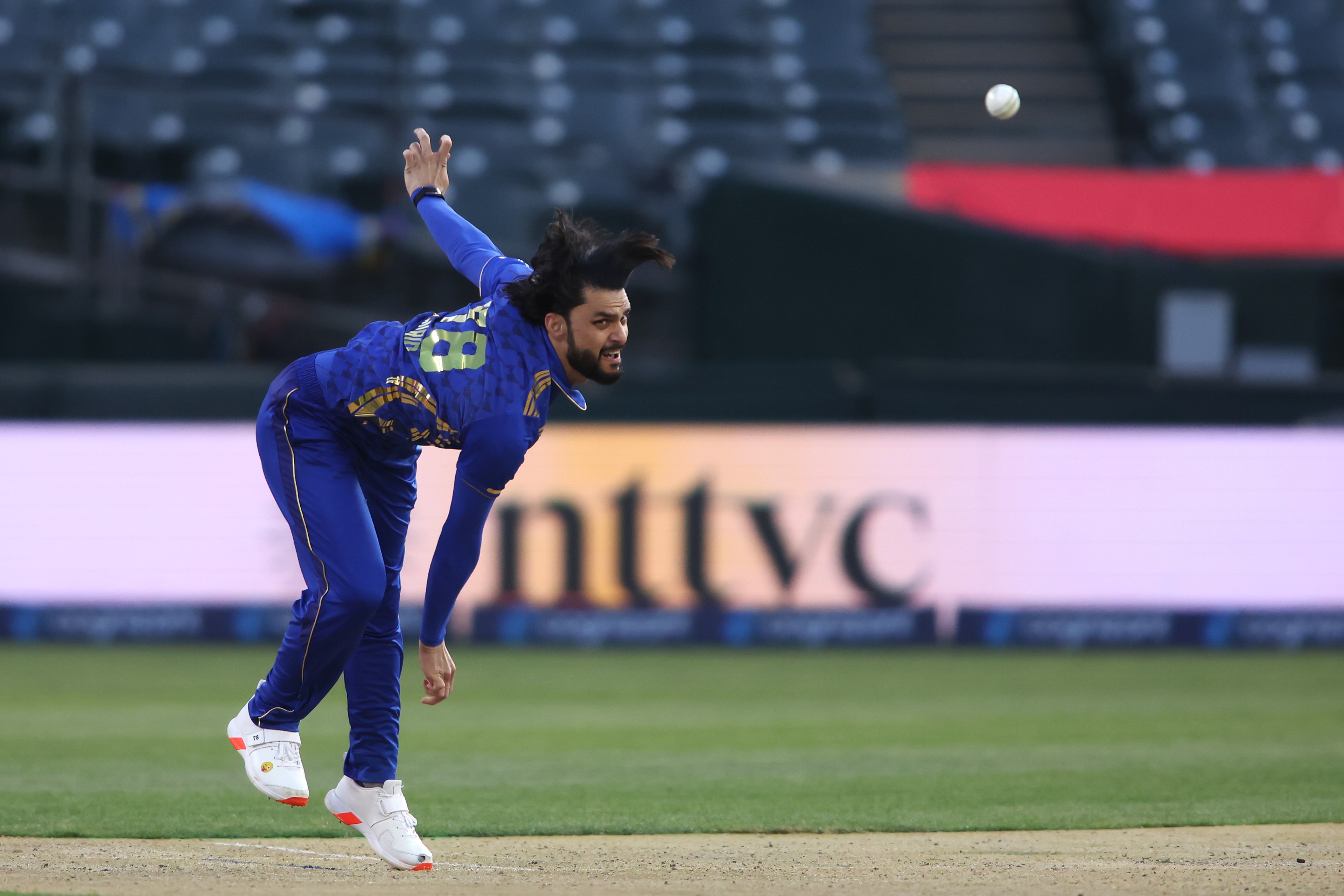 A cricket player in blue is bowling, mid-action, on a grassy field. The ball is in the air, and the background is a blurred stadium with empty seats.