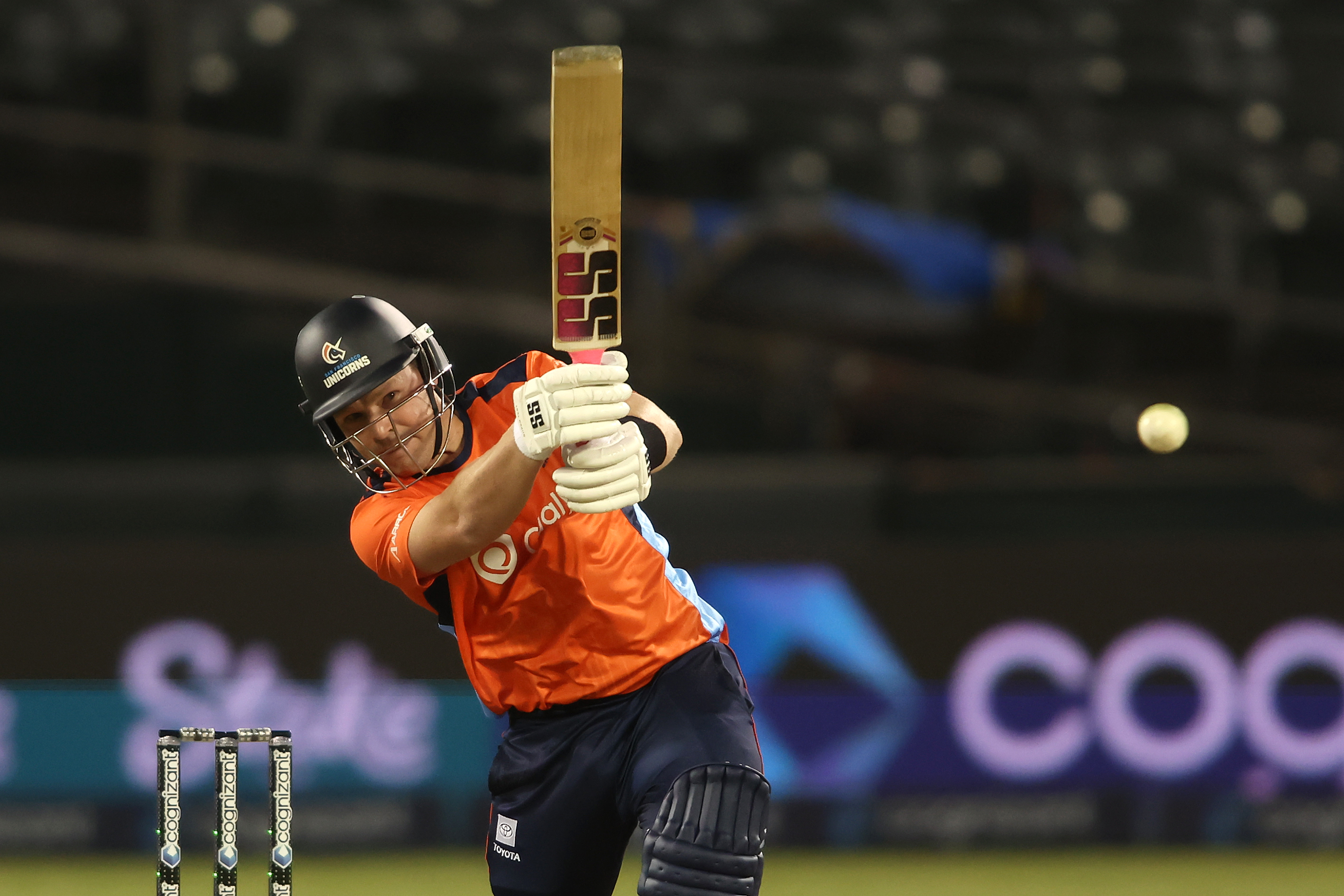 A cricket player in an orange jersey and helmet swings a bat, the ball is in mid-air. The background shows stadium seats and a blurred advertisement.