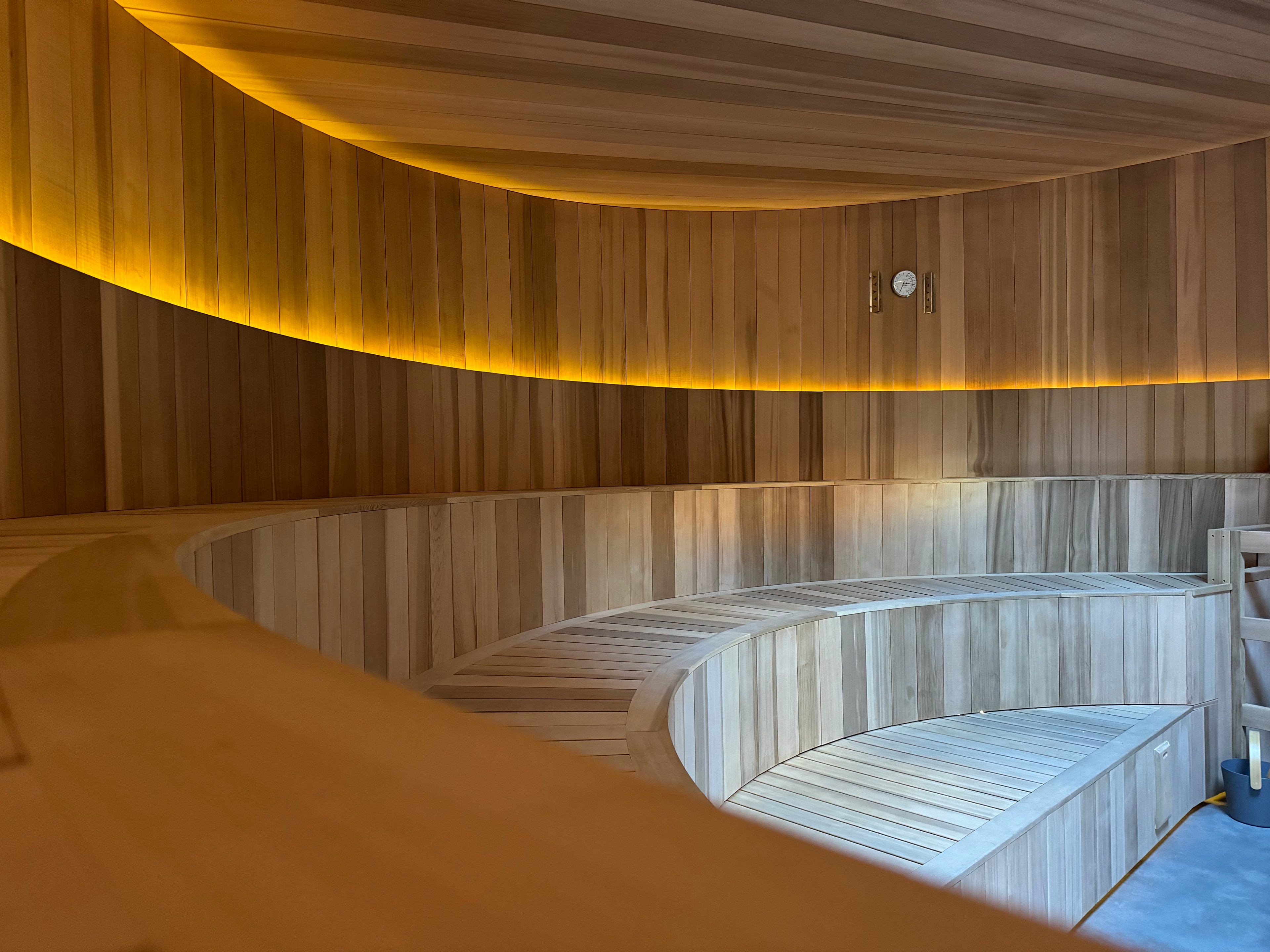 A wooden sauna with curved benches and warm, ambient lighting. A thermometer and hygrometer are mounted on the wall, adding to the cozy atmosphere.