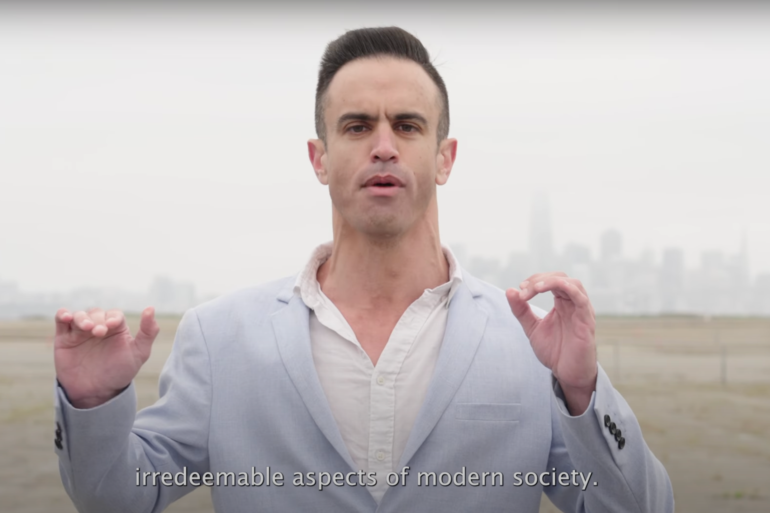 A person in a light blue suit stands outdoors with a city skyline in the background. Their hands are raised, and the text reads: "irredeemable aspects of modern society."