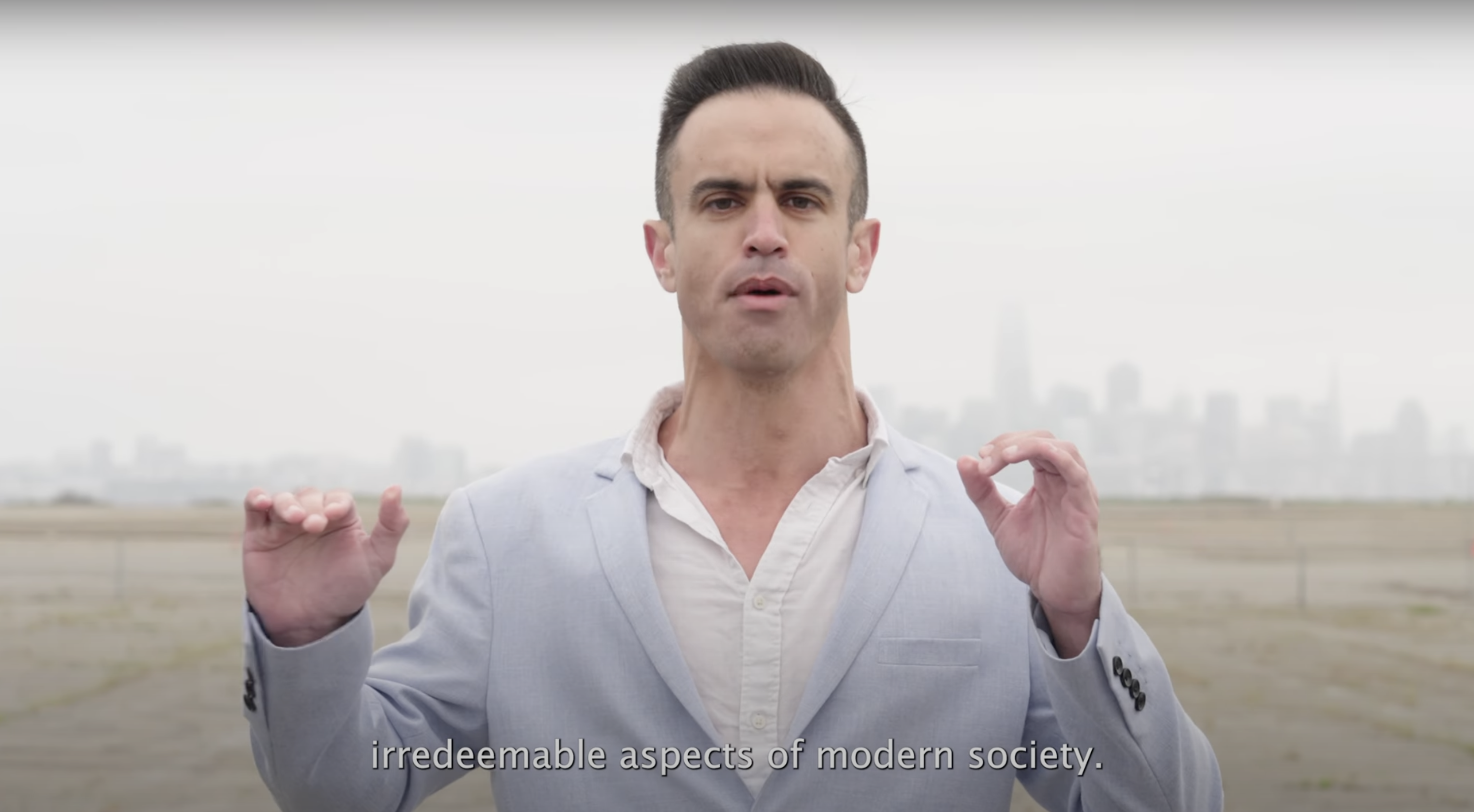 A person in a light blue suit stands outdoors with a city skyline in the background. Their hands are raised, and the text reads: "irredeemable aspects of modern society."