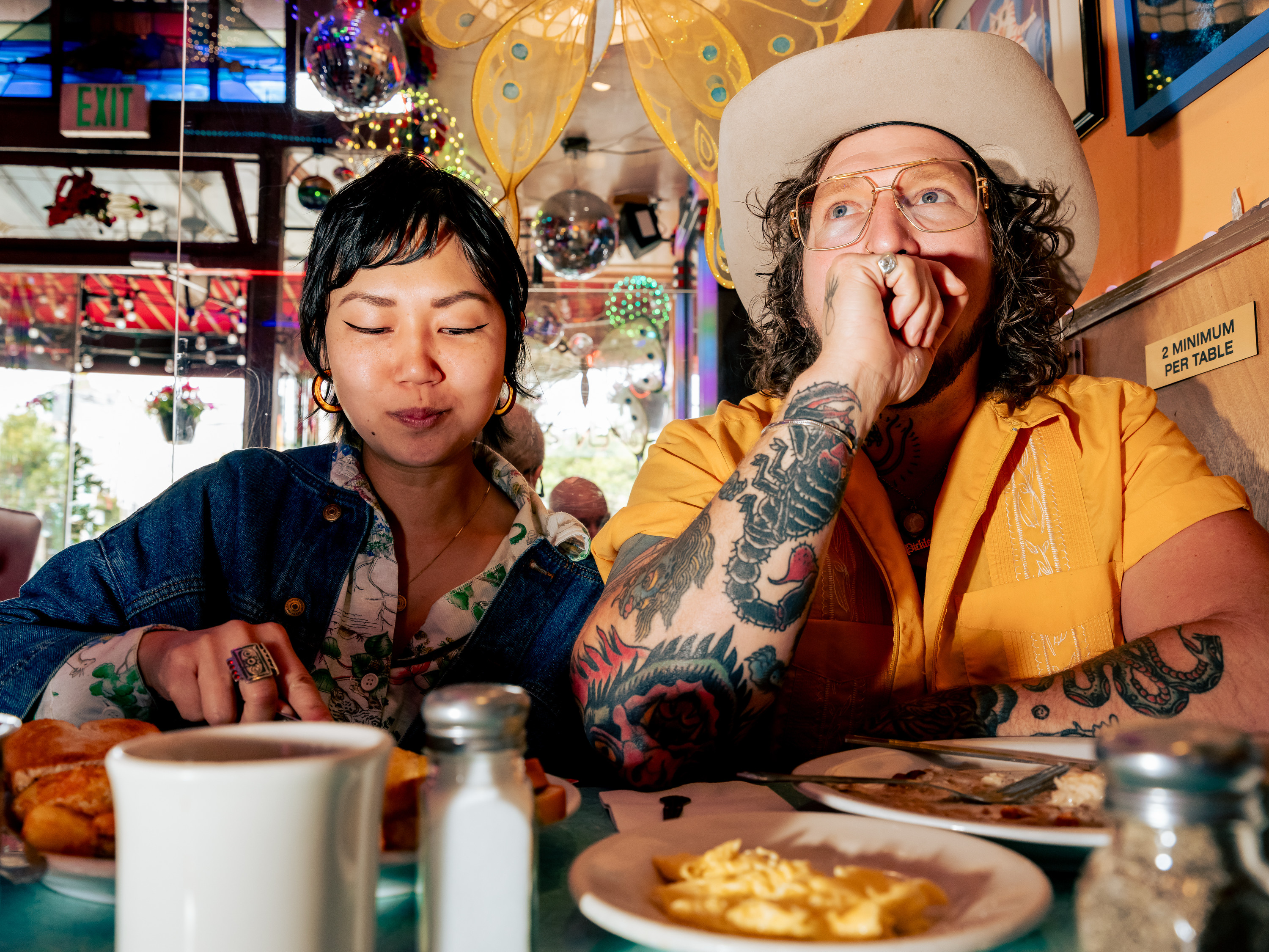 A woman and man sit at a colorful diner table with breakfast foods. She eats calmly; he, wearing a hat and tattoos, looks contemplative. Bright decor surrounds them.
