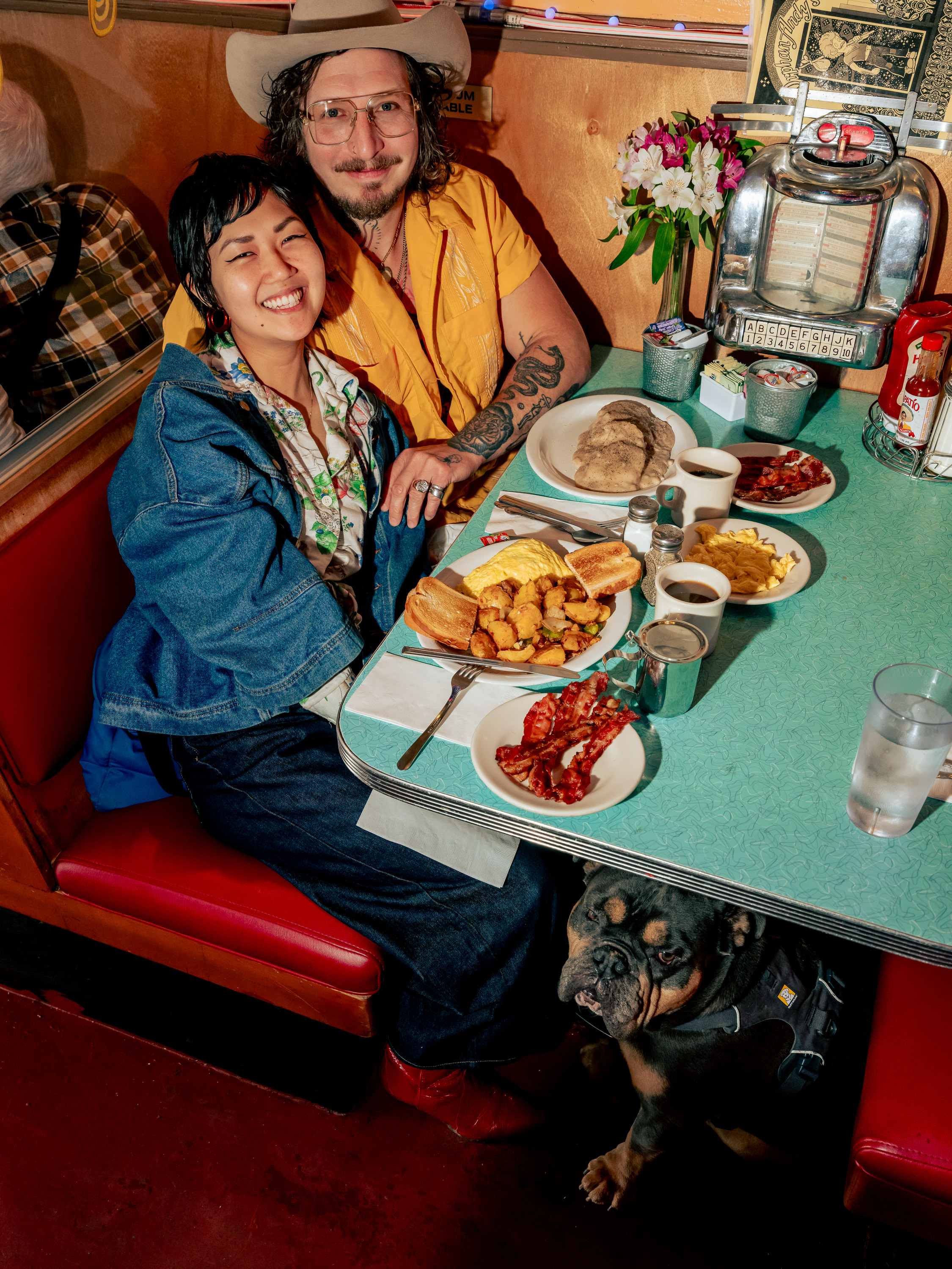 A smiling couple at a diner booth, surrounded by breakfast plates, with a relaxed dog sitting under the table. The booth has retro decor and flowers.
