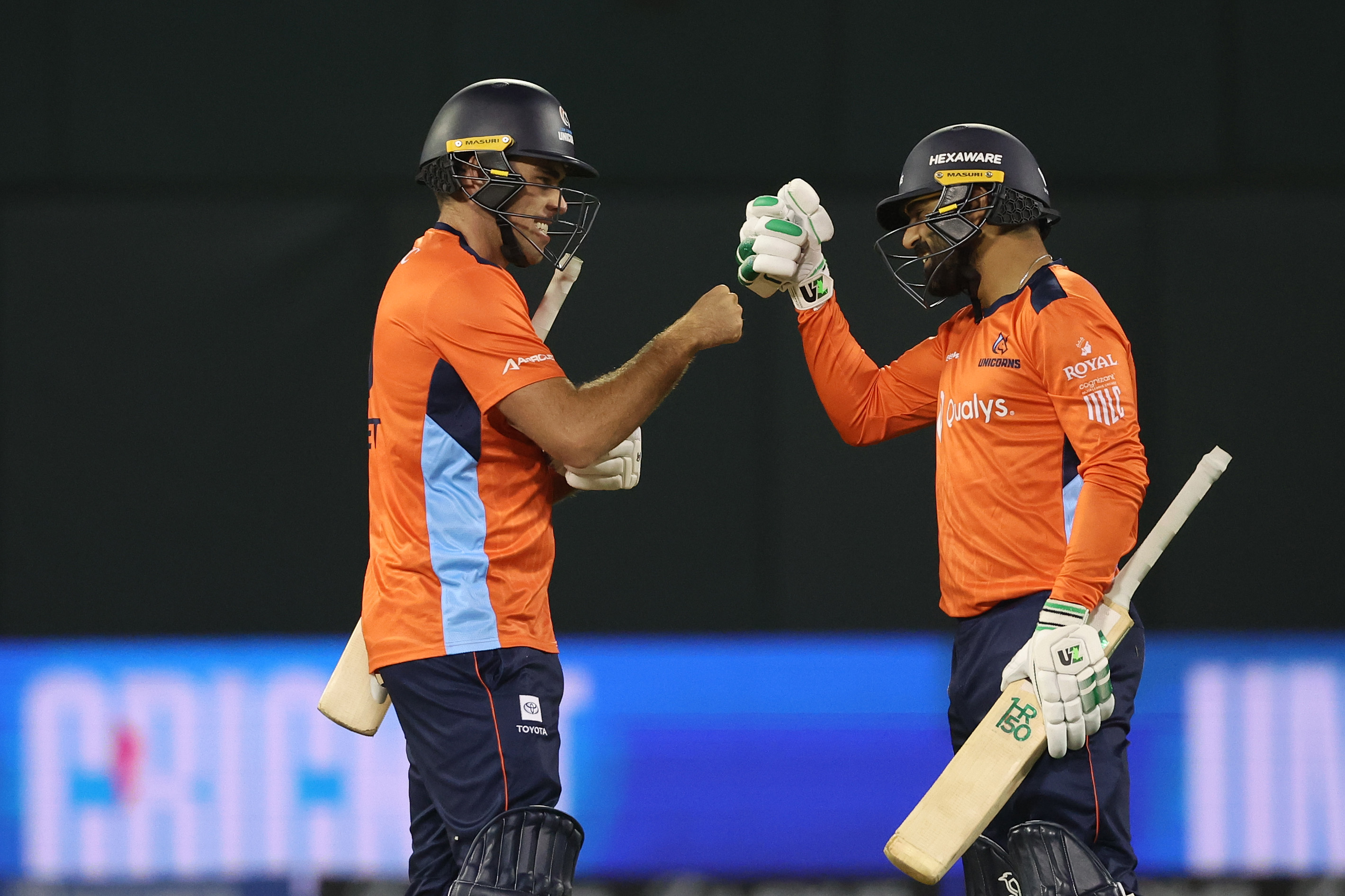Two cricketers in orange jerseys are fist-bumping on the field, both wearing helmets and holding bats, celebrating or strategizing.