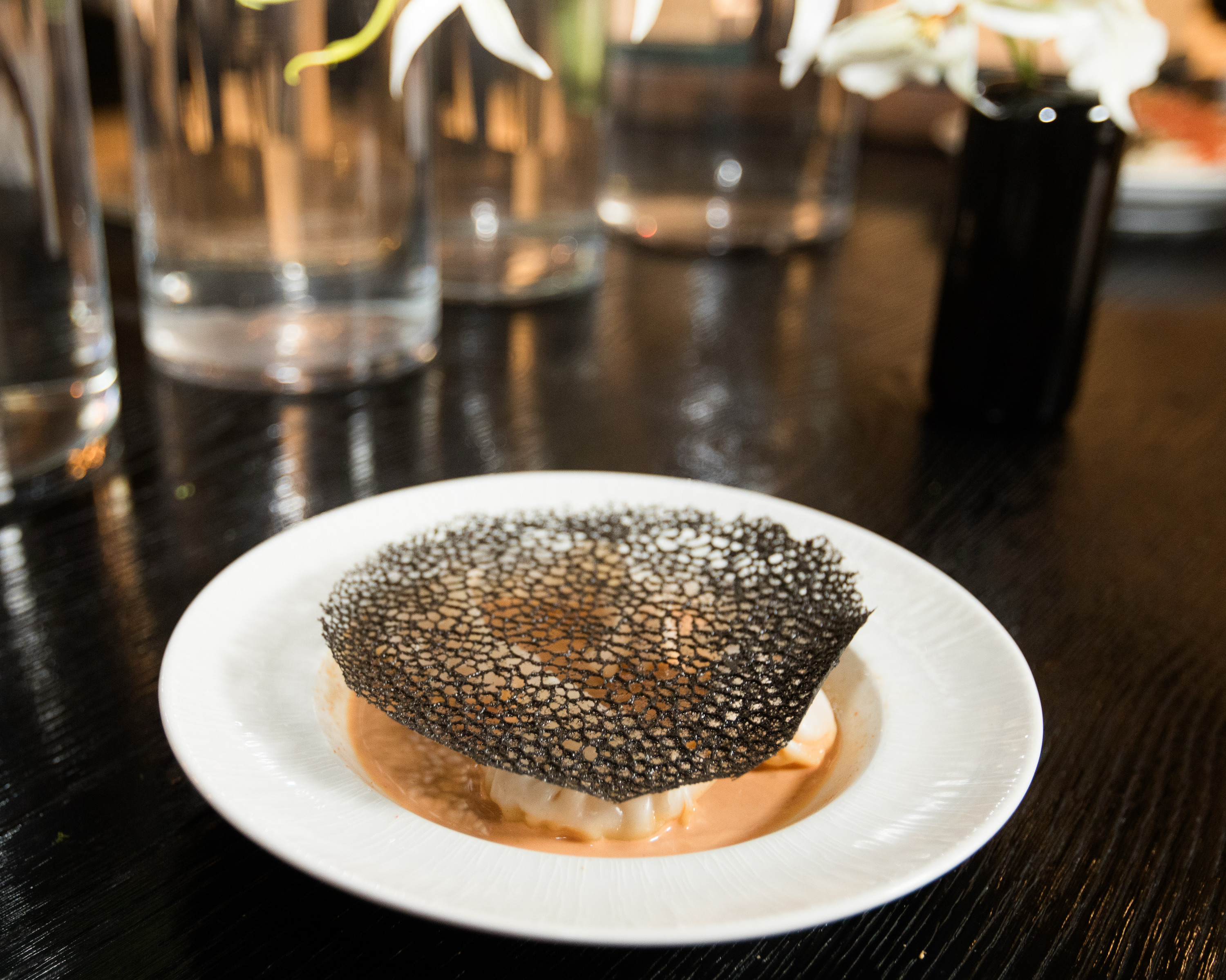 A white plate holds a dish topped with a delicate, black lace-like crisp on a creamy beige sauce. The background shows clear glass vases and a dark surface.
