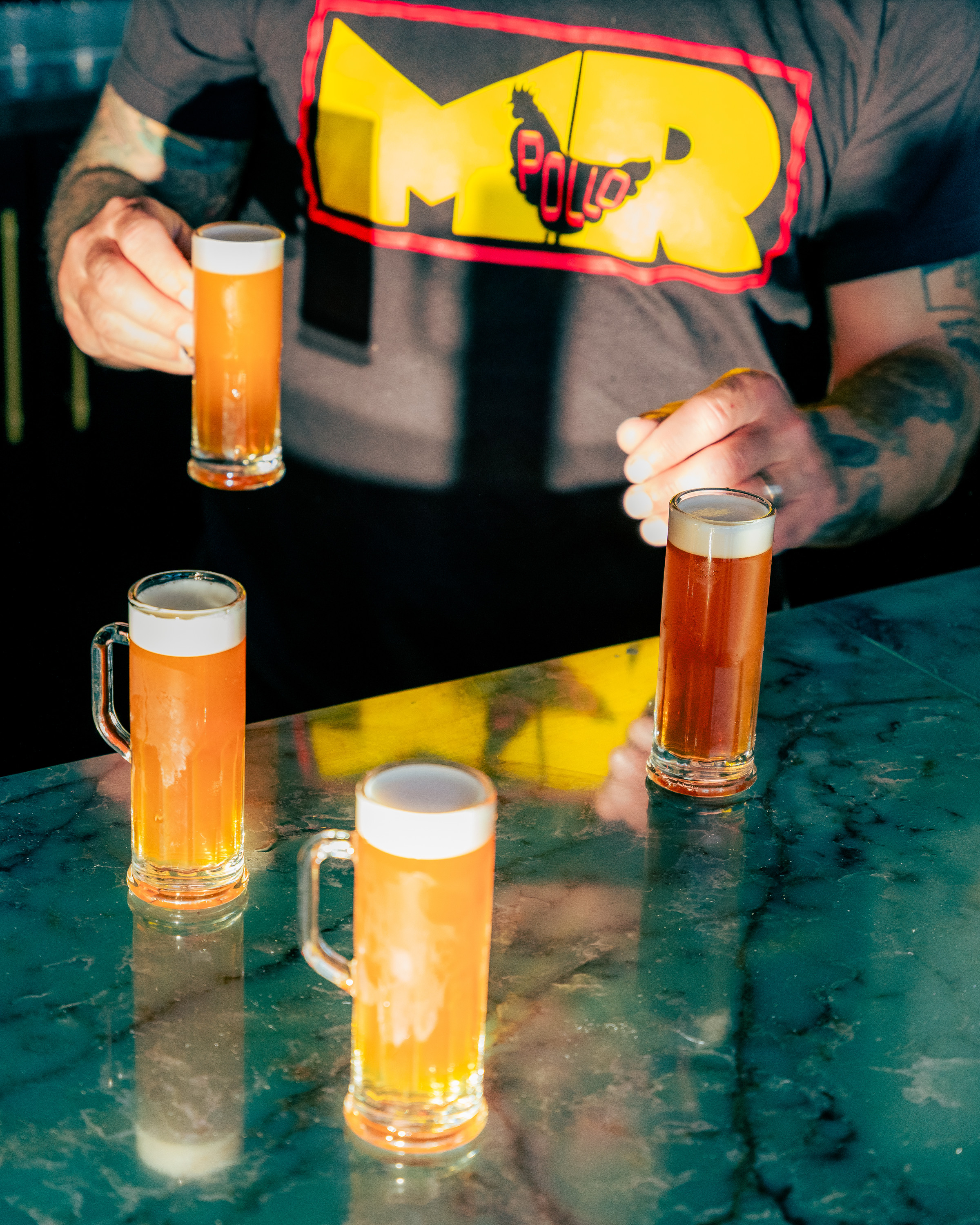A person is holding one of four frothy beer mugs on a marble surface, wearing a shirt with a bold yellow and red emblem with &quot;Pollo&quot; written on it.