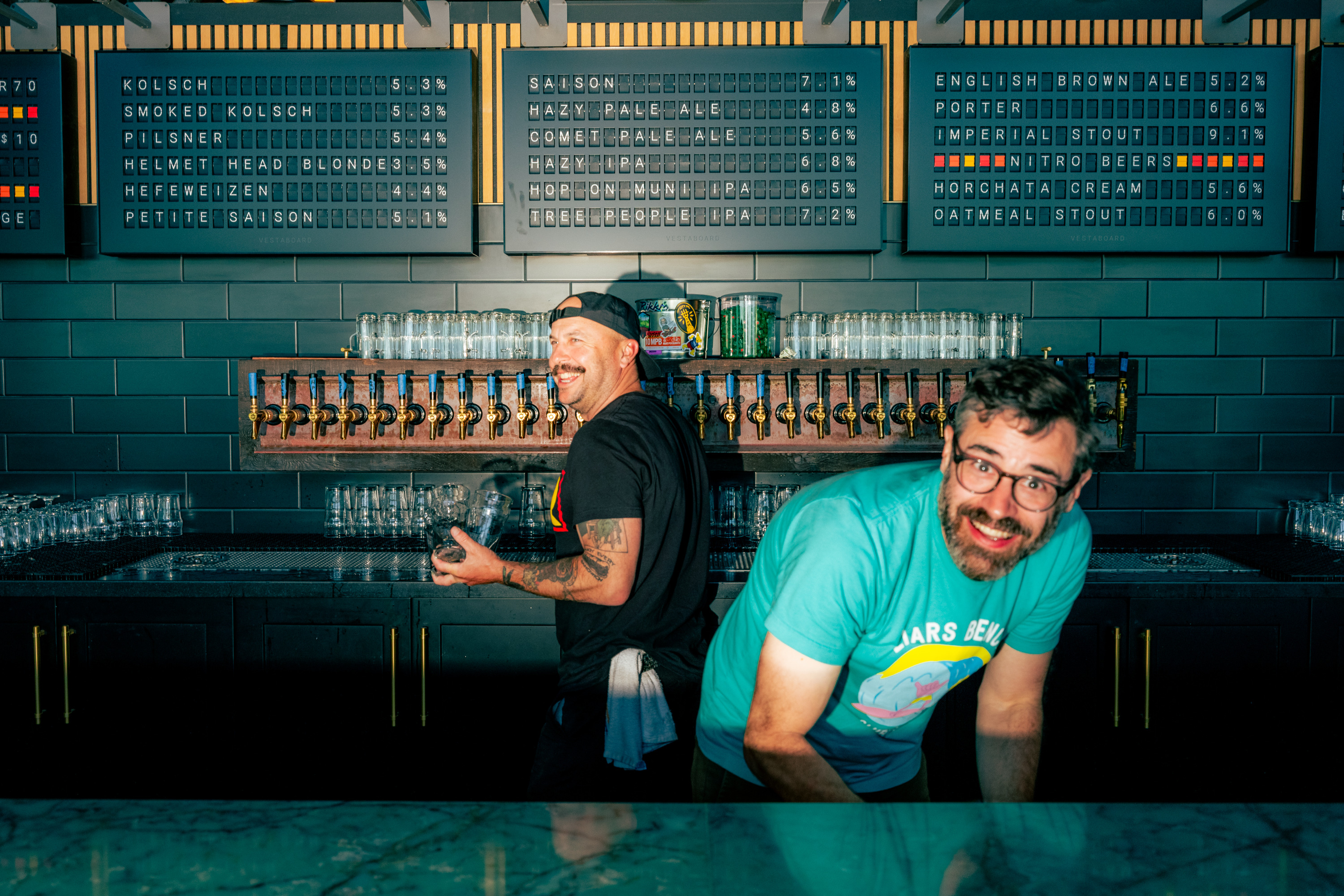 Two smiling men stand behind a bar with taps and glasses, surrounded by a menu board listing different beer types and alcohol percentages.