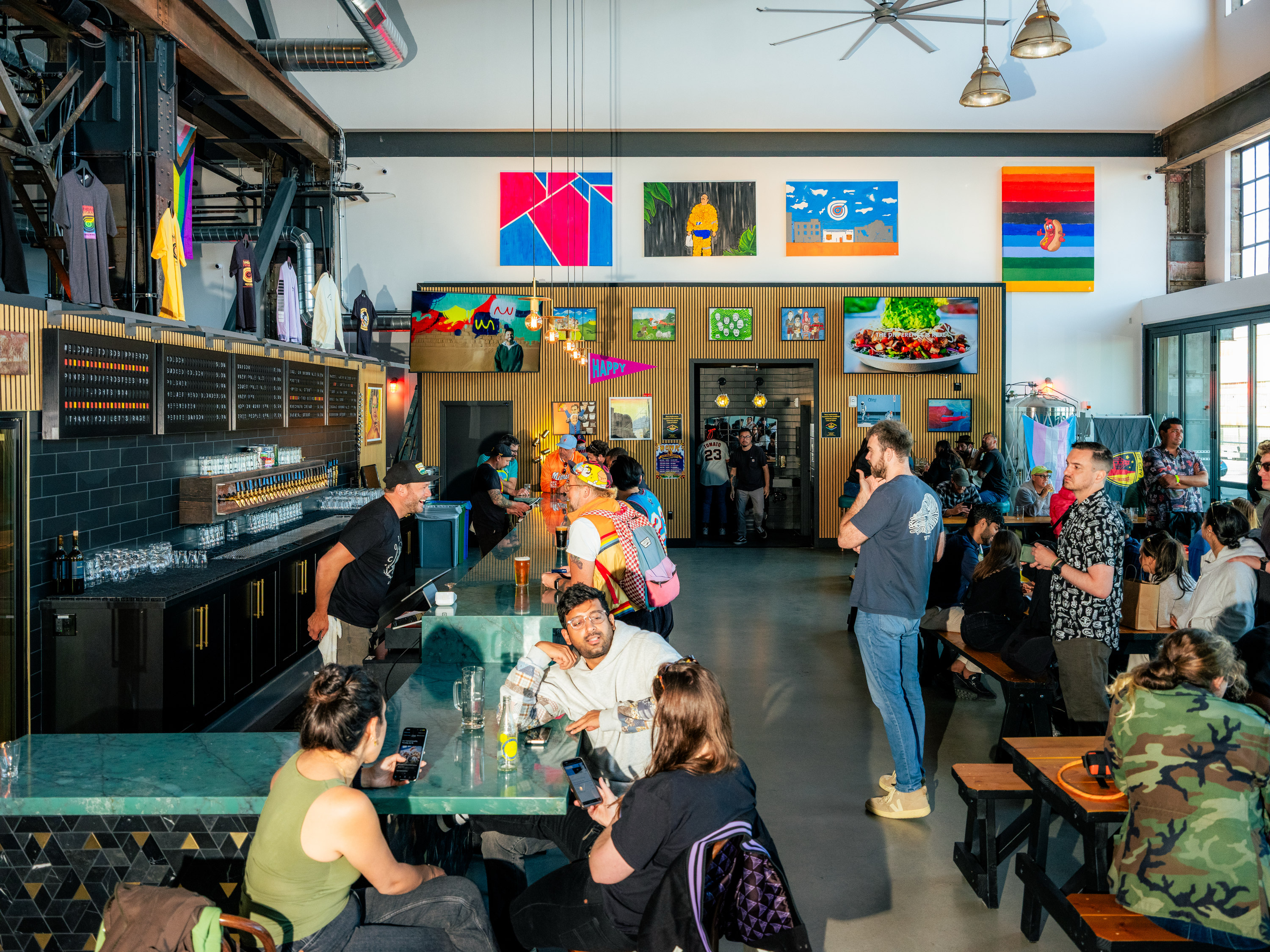 The image shows a lively bar with people sitting and chatting at tables. Art adorns the walls, and t-shirts hang above the bar. It's a bright, colorful space.