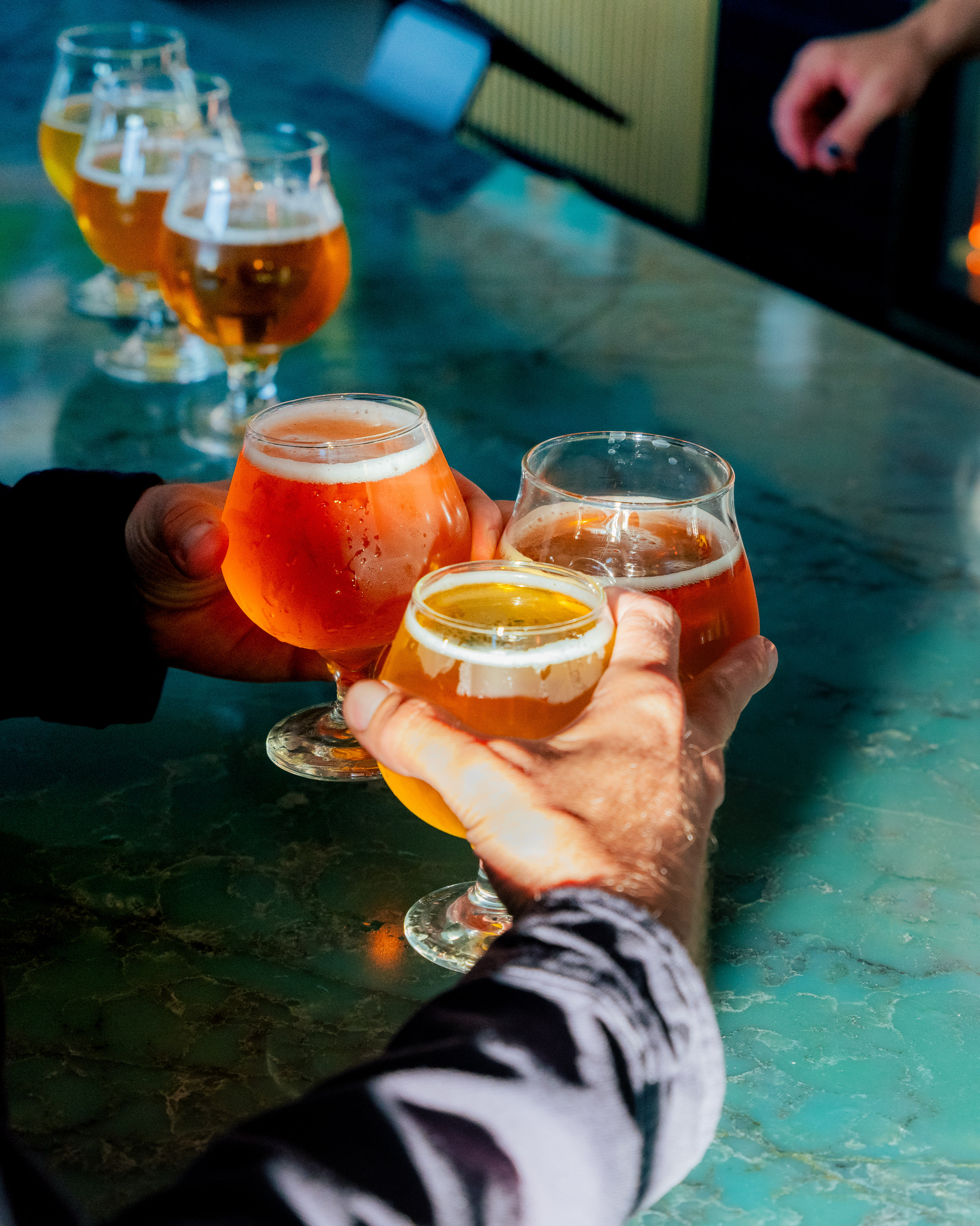 Several hands hold glasses of amber and golden beer, toasting over a green marble table in a well-lit bar setting.