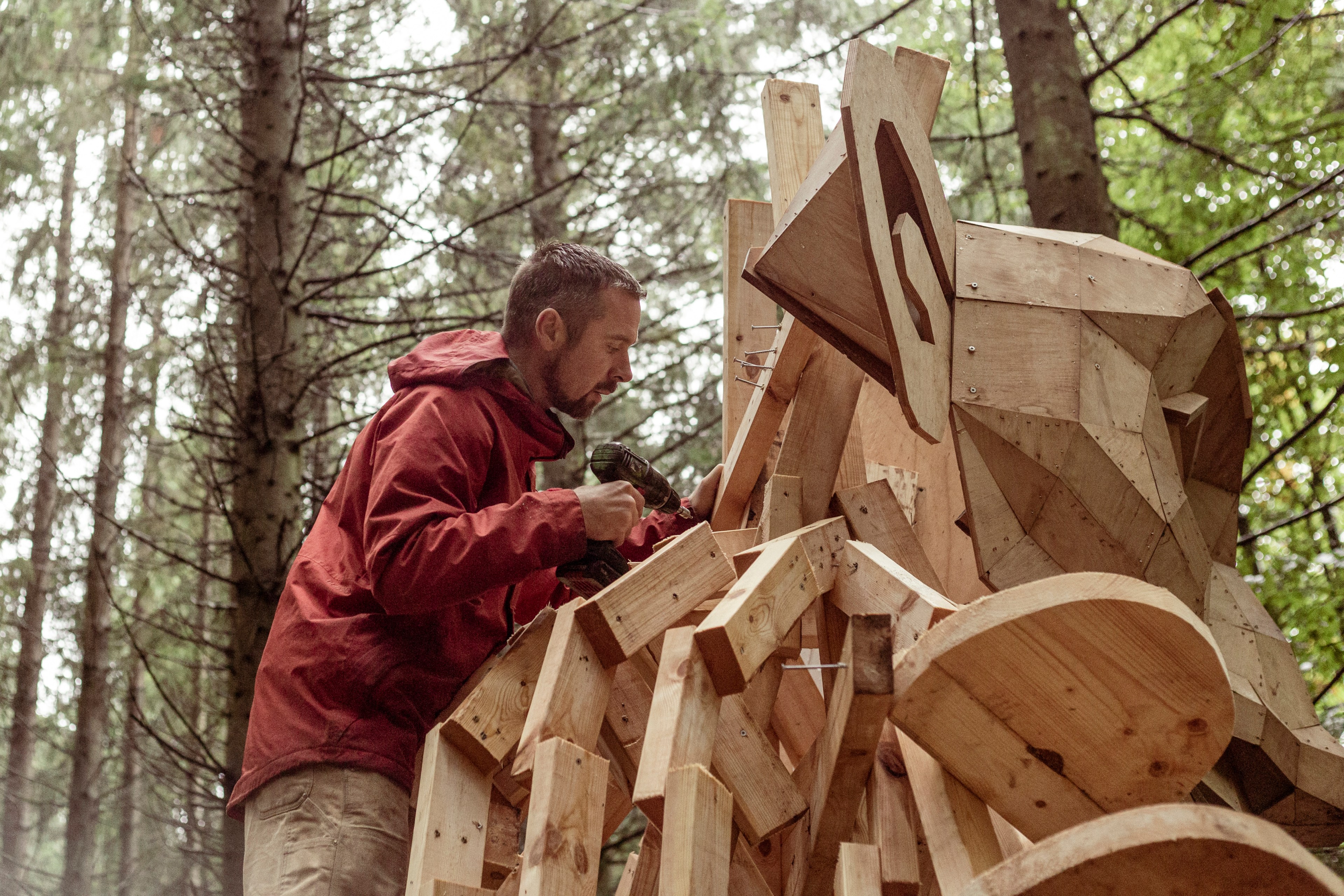 A person in a red jacket is using a power drill to work on a large, wooden sculpture in a forest. The sculpture has geometric shapes and resembles an animal.