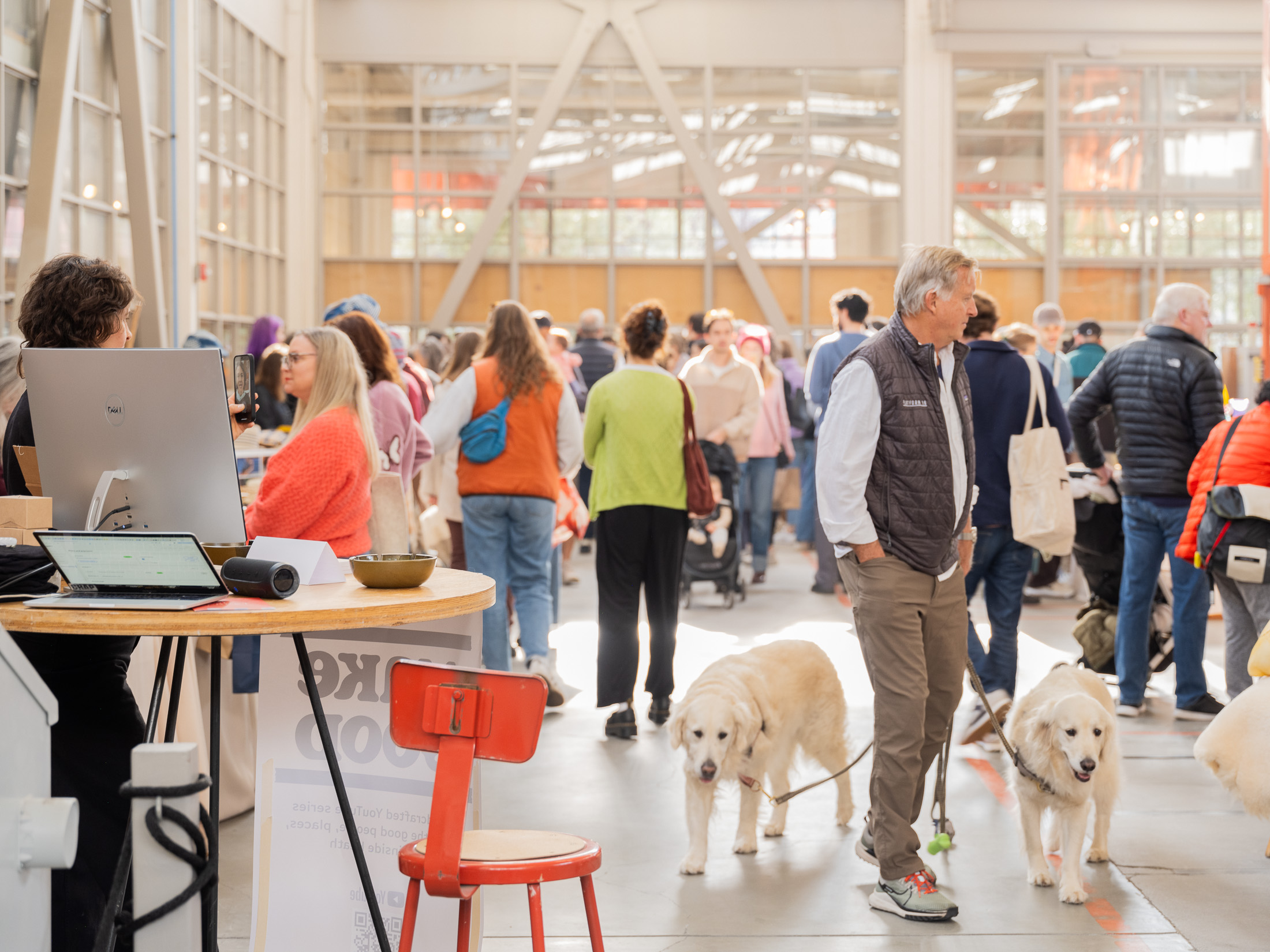 A bustling indoor market scene with people wearing casual clothing, some carrying bags, and a man walking two large white dogs on leashes.