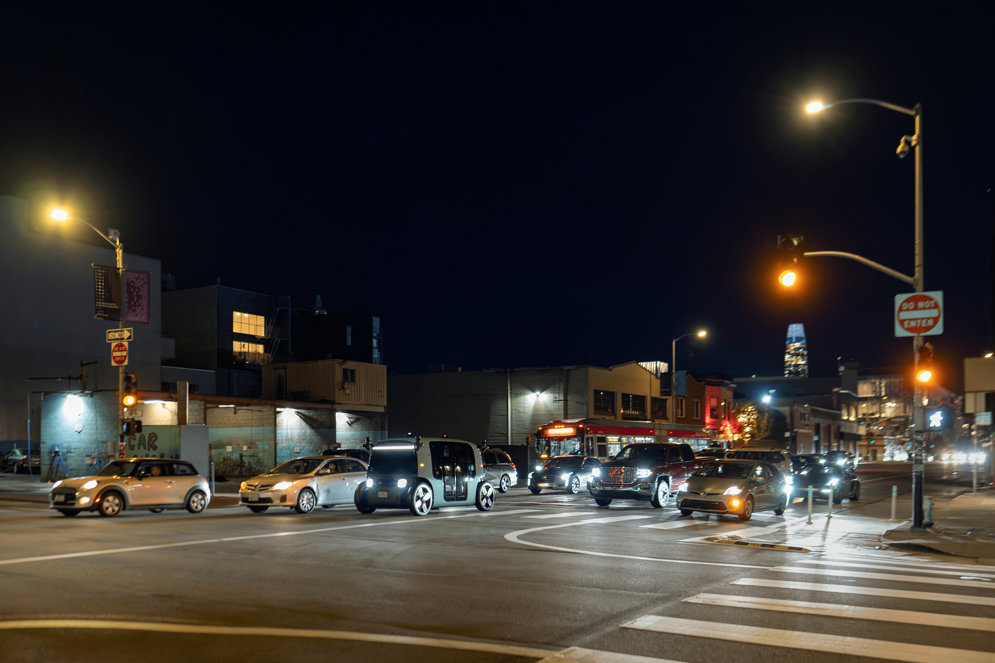 A nighttime urban scene features a busy intersection with cars stopped at a light, illuminated by streetlights, and city buildings in the background.