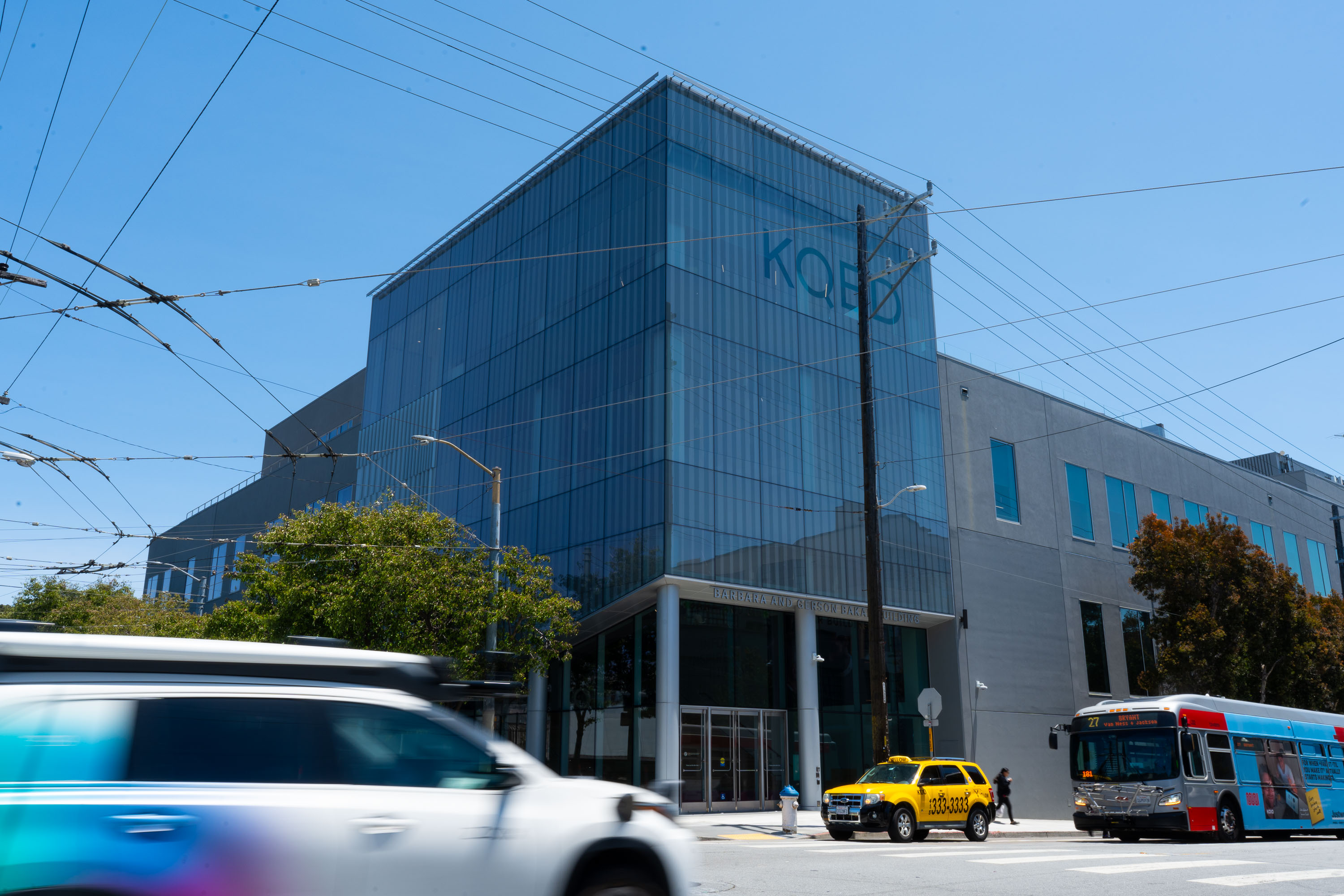 The image features a modern building with large glass windows labeled "KQED." Cars and a bus are passing by on the street, with overhead power lines visible.